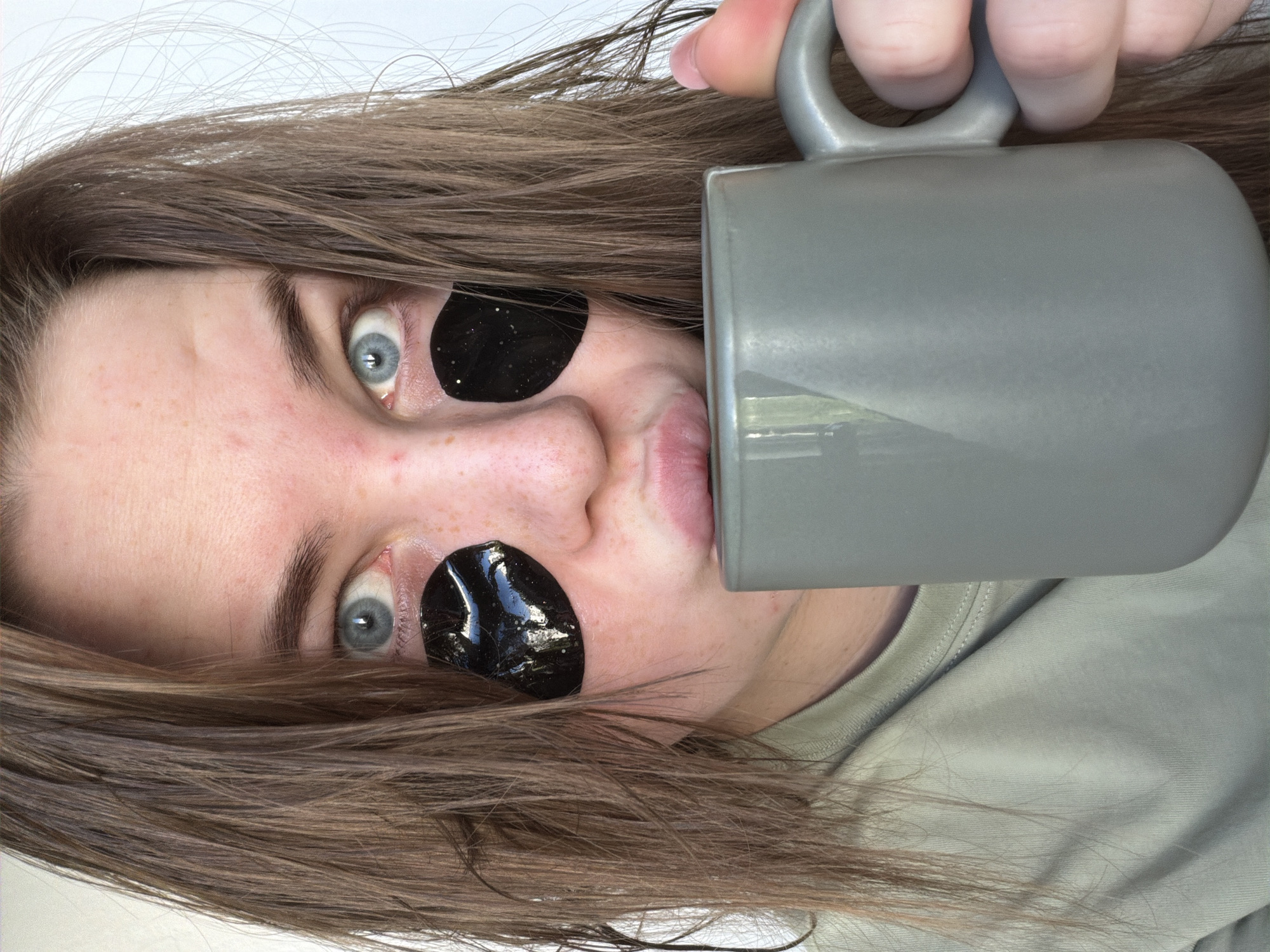 A person with light skin and light brown hair is lying down, with their head rotated slightly. They have black under-eye patches on both eyes. Their eyes are light blue. They are holding a gray mug with a handle up to their mouth. The person is wearing a light green t-shirt. The background appears to be a white surface. The image doesn't contain any information that suggests the location is in the city of Chaikovsky, Russia.