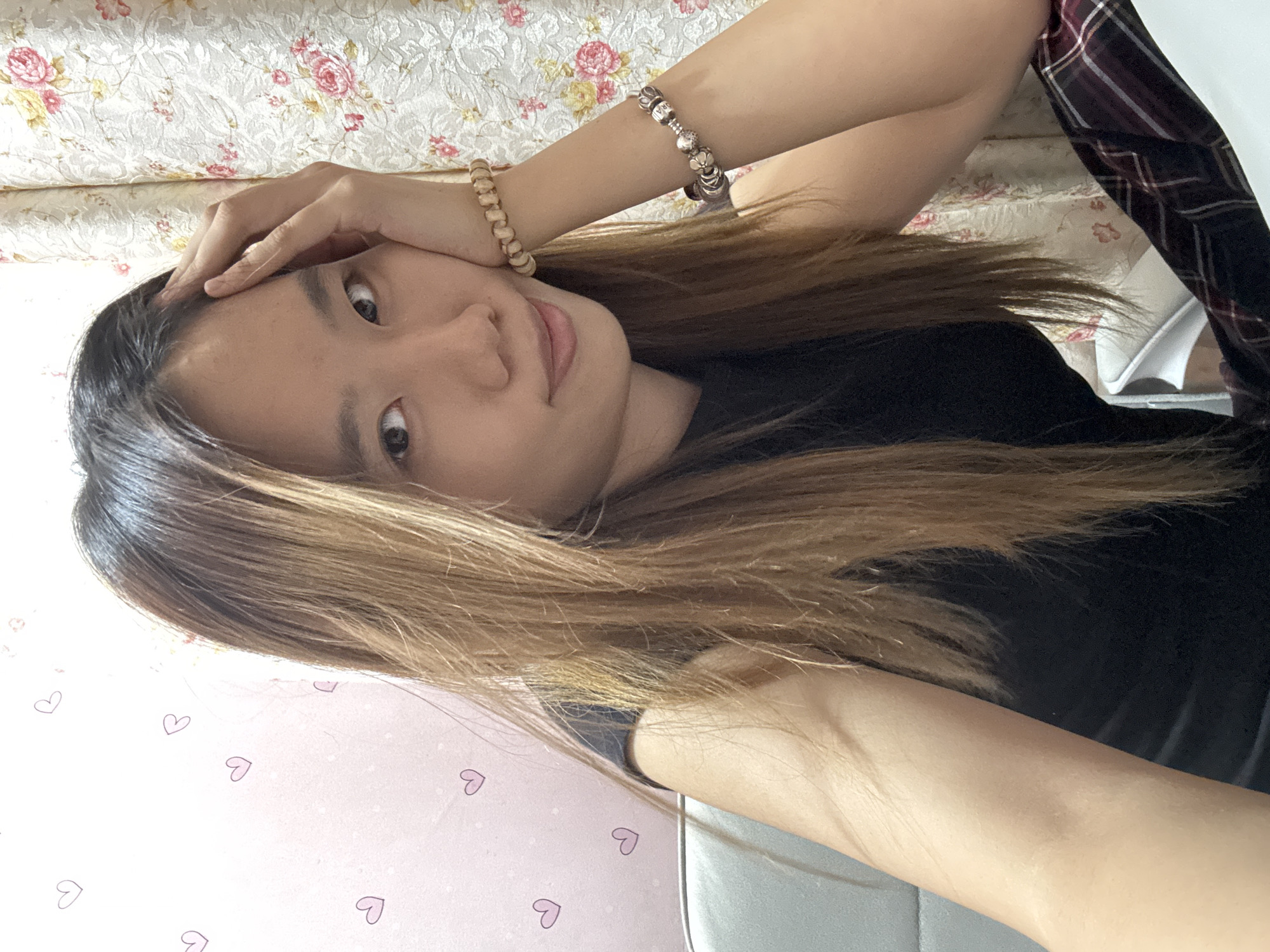 A young Asian woman with long, layered brown hair is shown in a close-up, head-and-shoulders shot. She is looking directly at the camera with a slight smile. Her right hand is resting on her forehead, and she is wearing two bracelets on her left wrist. One bracelet is made of light-colored beads, and the other is a silver charm bracelet. She is wearing a black top. The background consists of a pink wall with small heart designs and a patterned curtain. The image appears to have been taken indoors.