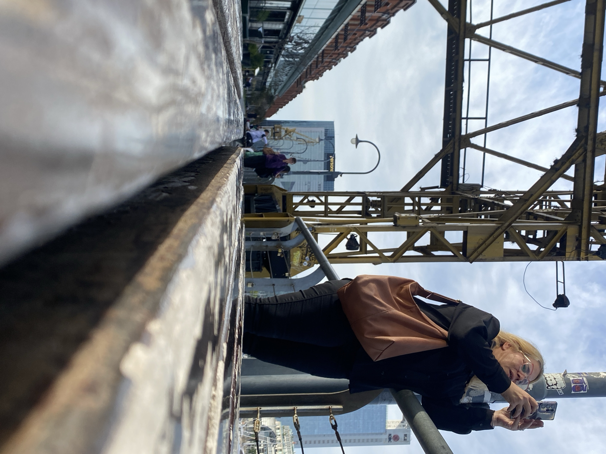 A medium, canted, outdoor shot shows a fair-skinned woman with blonde hair and glasses leaning forward while holding a smartphone in front of her to take a photograph, with a yellow crane extending above her to the right. The woman is wearing a black buttoned shirt and pants, and a light-brown leather handbag is slung over her shoulder. She is standing on a structure of metal poles with decorative cables hanging down in the background, presumably a bridge or overpass.
Behind the crane, to the upper left, is a building with “PERSONE” written vertically on its side, and behind that are pedestrians walking along the building's side. Above the pedestrians, other buildings extend toward the left of the shot. The sky is cloudy and visible through the crane's structure. A tall white building with a red logo can be seen to the left of the woman. The foreground of the shot has a white ledge or railing with gray streaking.