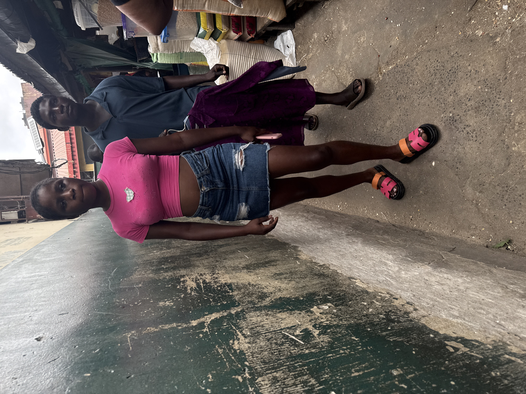 The image shows a young woman and a young man standing in an outdoor market in Lagos, Nigeria. The woman is in the foreground, wearing a pink crop top and a denim skirt. She is looking towards the camera with a neutral expression. The man is behind her, wearing a blue polo shirt and holding a purple garment. They appear to be walking together through a crowded market, with stalls selling various goods visible in the background. The environment is an urban street scene, with a textured wall on the left and a pathway on the right. The overall lighting suggests it is daytime, possibly overcast.