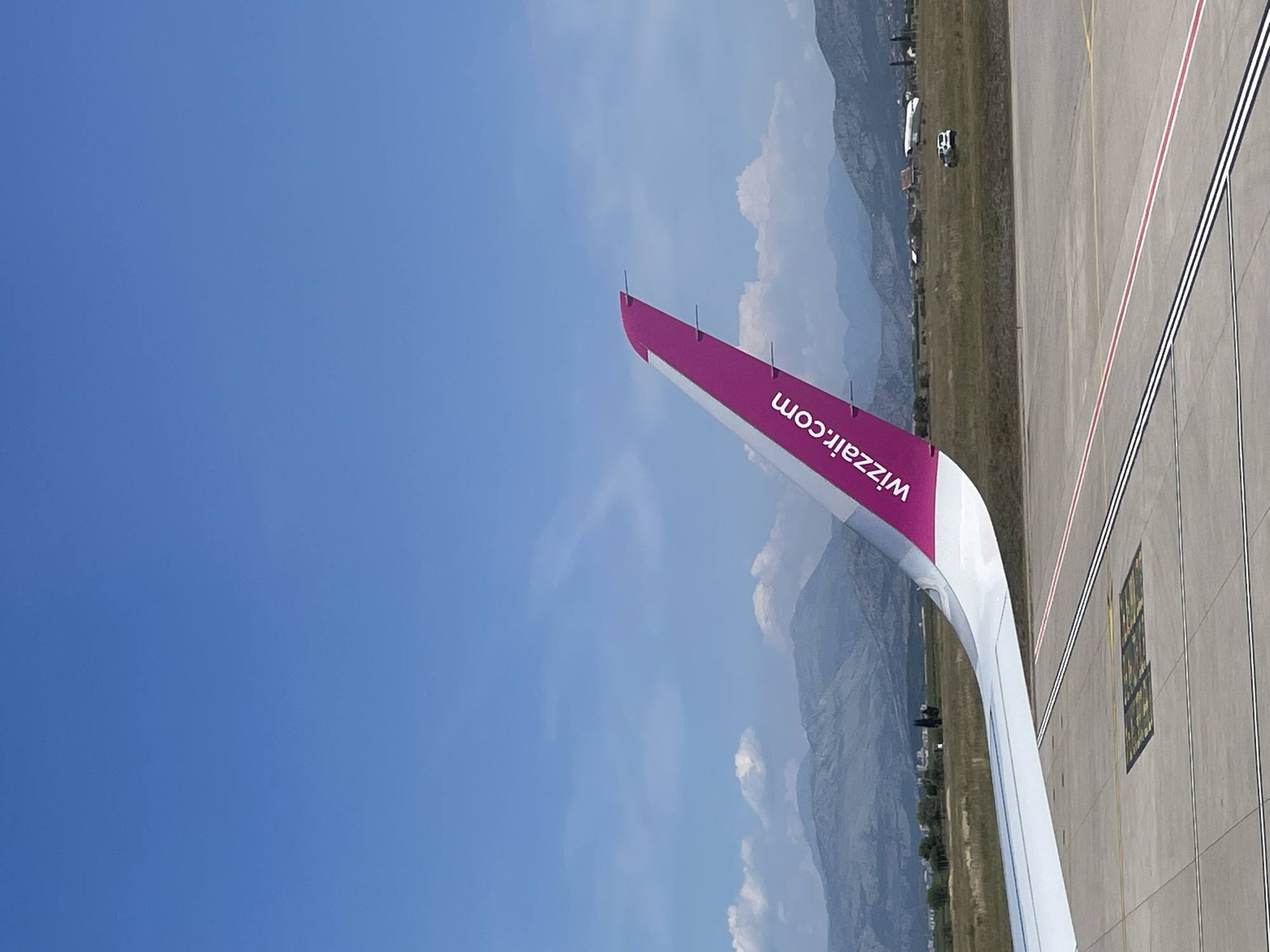 The image shows the tail of a Wizz Air airplane at Podgorica Airport in Montenegro. The tail is painted with the Wizz Air logo and website address (wizzair.com). The background shows the airport tarmac, green grassy areas, mountains, and a partly cloudy sky. Some airport buildings are also visible in the distance.