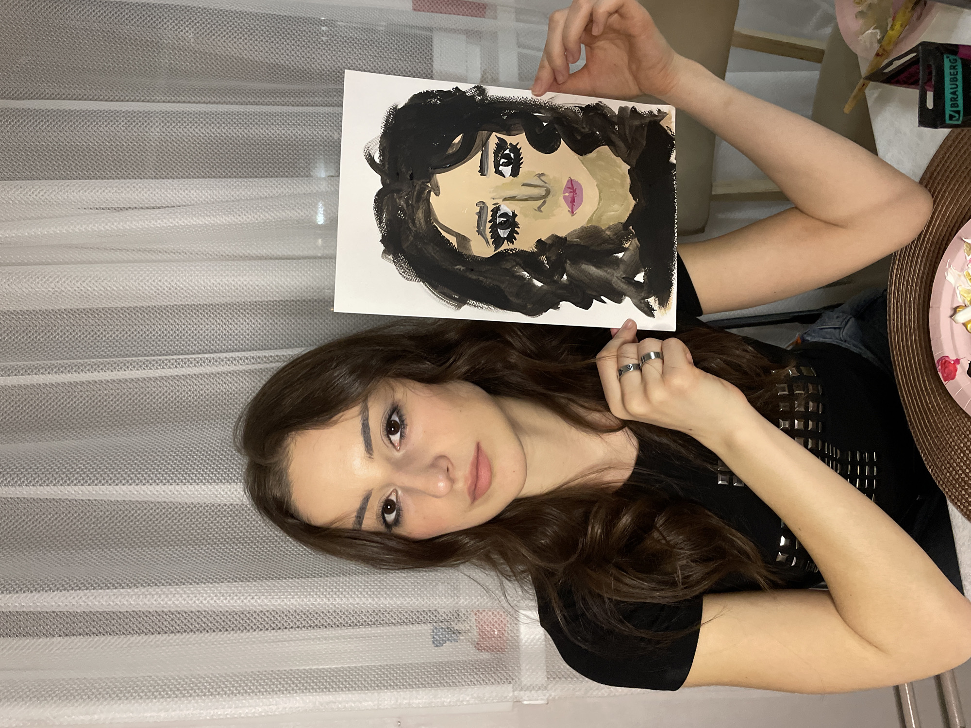 A young woman with long, dark brown hair and dark eyes is depicted indoors, holding up a painting she created. She looks directly at the camera with a calm, neutral expression. She is wearing a black top adorned with metallic studs and several silver rings on her left hand.
The painting she holds is a stylized portrait of a face, characterized by broad, expressive brushstrokes. It features dark, wavy hair, light skin, exaggerated dark eyes with prominent black eyeliner or mascara, and distinct pink lips. The art appears to be executed in an expressive style, possibly using acrylics or gouache.
The background shows sheer white curtains with horizontal grey mesh stripes, suggesting an indoor setting, likely a home studio or living space. To the right, a table with a woven brown placemat holds art supplies, including a small pink dish with various paint colors (white, yellow, red) and a black container with the brand name "BRAUBERG" visible. The lighting is consistent with an indoor setting, possibly during the evening. The scene takes place in Khimki, Russia.