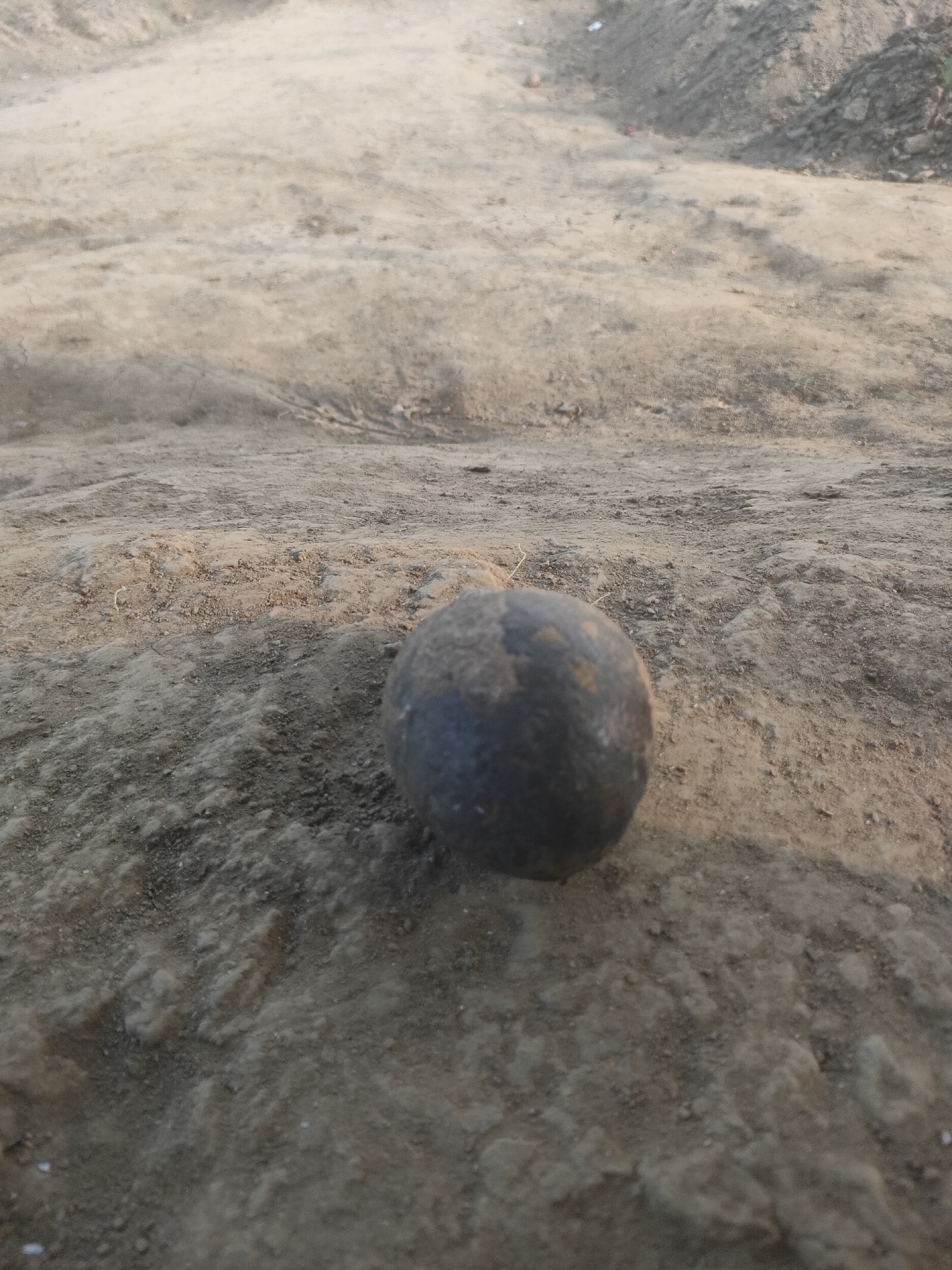 The image displays a dark, spherical object resting on a patch of dry, sandy soil. The object, roughly the size of a fist, appears to be a cannonball or a similar heavy projectile. Its surface is textured and somewhat worn, with visible pitting and variations in color, suggesting age and exposure to the elements. The surrounding environment is predominantly barren, characterized by loose earth, small stones, and subtle undulations in the terrain. In the background, a sloped area of piled earth indicates a possible construction site or excavation. The lighting suggests it is either early morning or late afternoon, casting soft shadows and highlighting the rough texture of the ground. There are no people or any other discernible living organisms present in the scene. The overall impression is one of an outdoor, undeveloped area, possibly in a rural or semi-urban setting. This scene is located in the city of Padang Dalom, Indonesia.