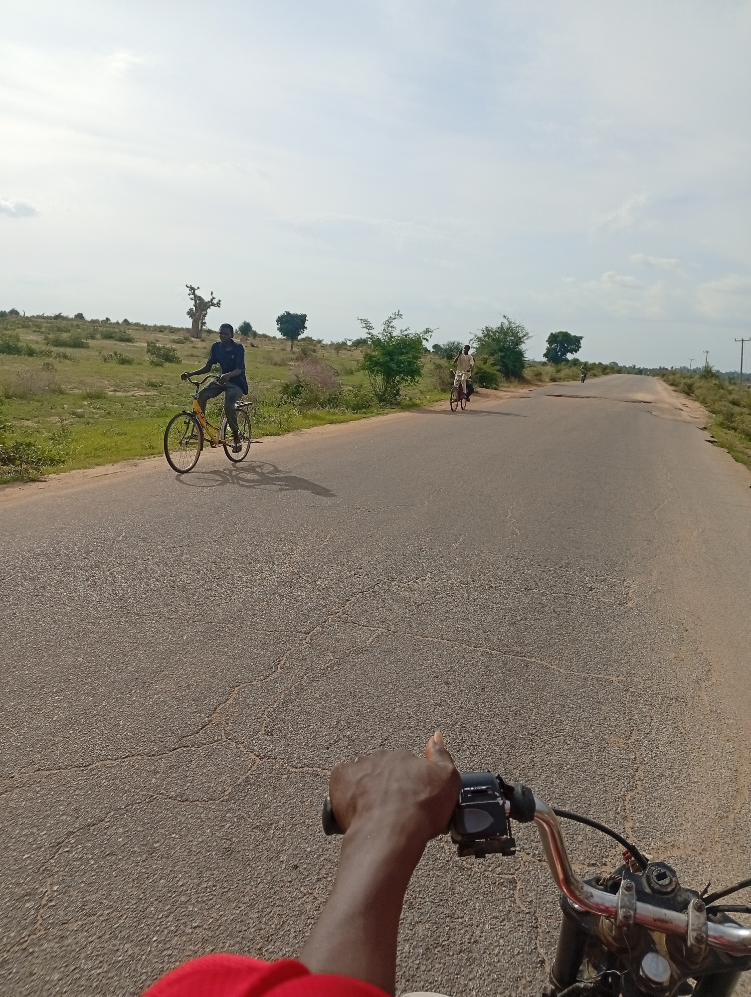 In Shuwa, Nigeria, this image captures a view from a motorcycle moving along an asphalt road. Two individuals on bicycles are visible in the distance on the same road. The landscape bordering the road is characterized by dry grass, scattered bushes, and a few trees, including a distinctive baobab tree. The sky is partly cloudy, with the sun casting shadows on the road. The focus is on the road ahead and the handlebars of the motorcycle, with a hand on the right side giving a thumbs-up gesture.