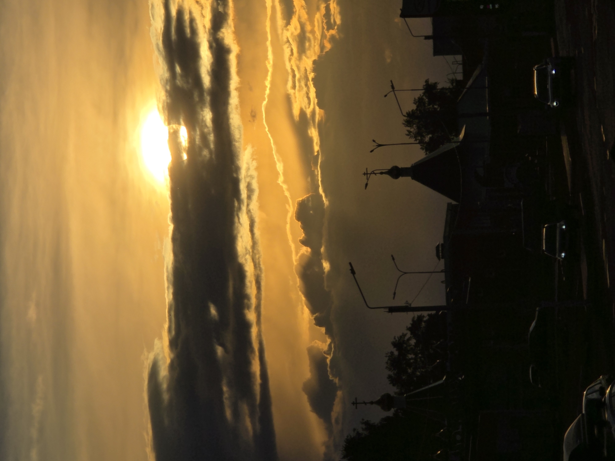 The image depicts a vertical view of a sunset scene in what appears to be Chaikovsky, Russia. The sun is partially obscured by dark, striated clouds on the left side of the frame, casting an orange glow across the sky and other clouds. In silhouette against the bright sky, there are structures that suggest a cityscape. These include the outline of a church with a prominent cross on top of its dome and buildings and streetlights. A vehicle is visible along a road. The lighting suggests a time near sunset.