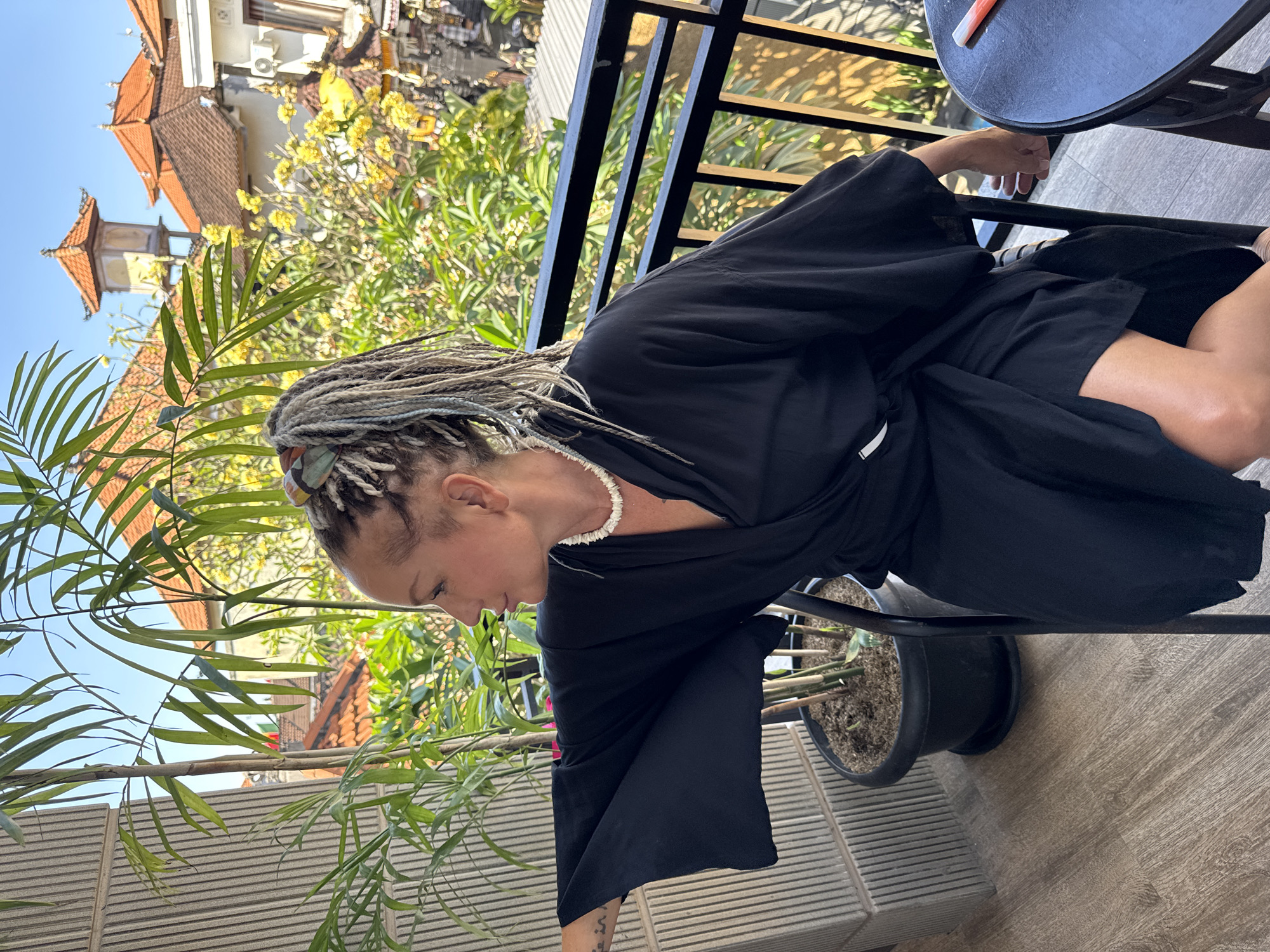 A woman with light brown dreadlocks is seated on a chair on a balcony. She is wearing a black robe and a white beaded necklace. The balcony overlooks a street with buildings, trees, and a clear blue sky. She is looking to her left. A black round table is to her right, with a red and white object on it. A potted plant is to her left. The floor of the balcony is made of wooden planks. The buildings in the background have orange tiled roofs and intricate architectural details, typical of Kuta, Indonesia.