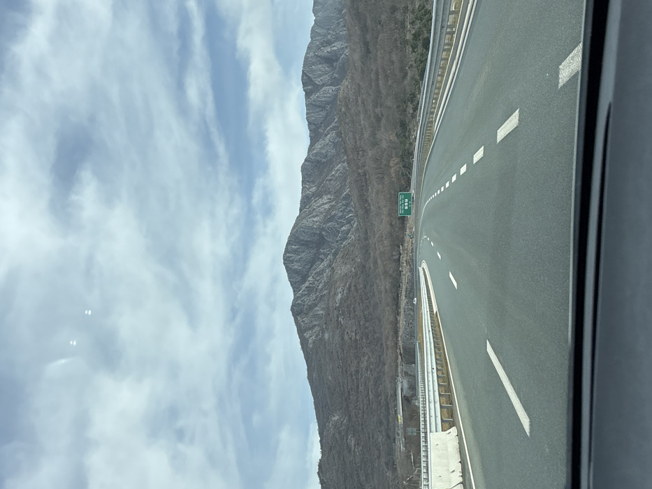 This image presents a forensic view of a highway segment near Pelev Brijeg, Montenegro, observed from within a vehicle. The asphalt highway features two lanes of travel in the foreground, demarcated by a solid white line on the left shoulder and dashed white lane separators, curving gently to the right. White concrete barriers line the left side, transitioning to metal guardrails further along, with additional metal guardrails lining the right side.
In the mid-ground, a green overhead road sign is visible, displaying destinations such as "Bioče," "Veternik," and "Podgorica" with associated distances, in both Cyrillic and Latin script. A partial bridge or overpass structure is visible to the left, beyond the concrete barriers. Minimal vehicle traffic, appearing as small white forms, is discernible in the distant lanes.
The background is dominated by a large, rugged mountain range characterized by prominent grey rock formations and slopes covered in sparse, bare deciduous vegetation, indicating a late autumn or winter season. The sky above is partly cloudy, exhibiting a mix of light blue and grey cloud formations. The scene is illuminated by natural daylight.