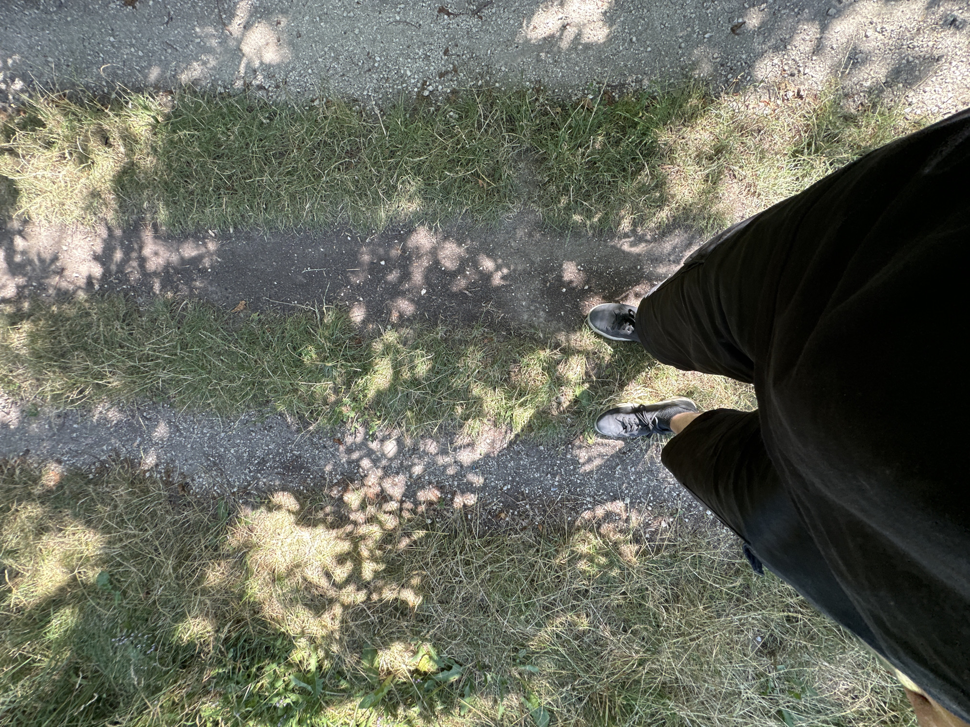 The image shows a person's legs and feet from a first-person perspective, standing on a dirt and gravel path bordered by dry grass. The path is dappled with shadows from overhead foliage, suggesting an outdoor, possibly wooded or park-like, environment. The person is wearing black shorts and grey athletic shoes. The location is identified as Makiv, Ukraine.