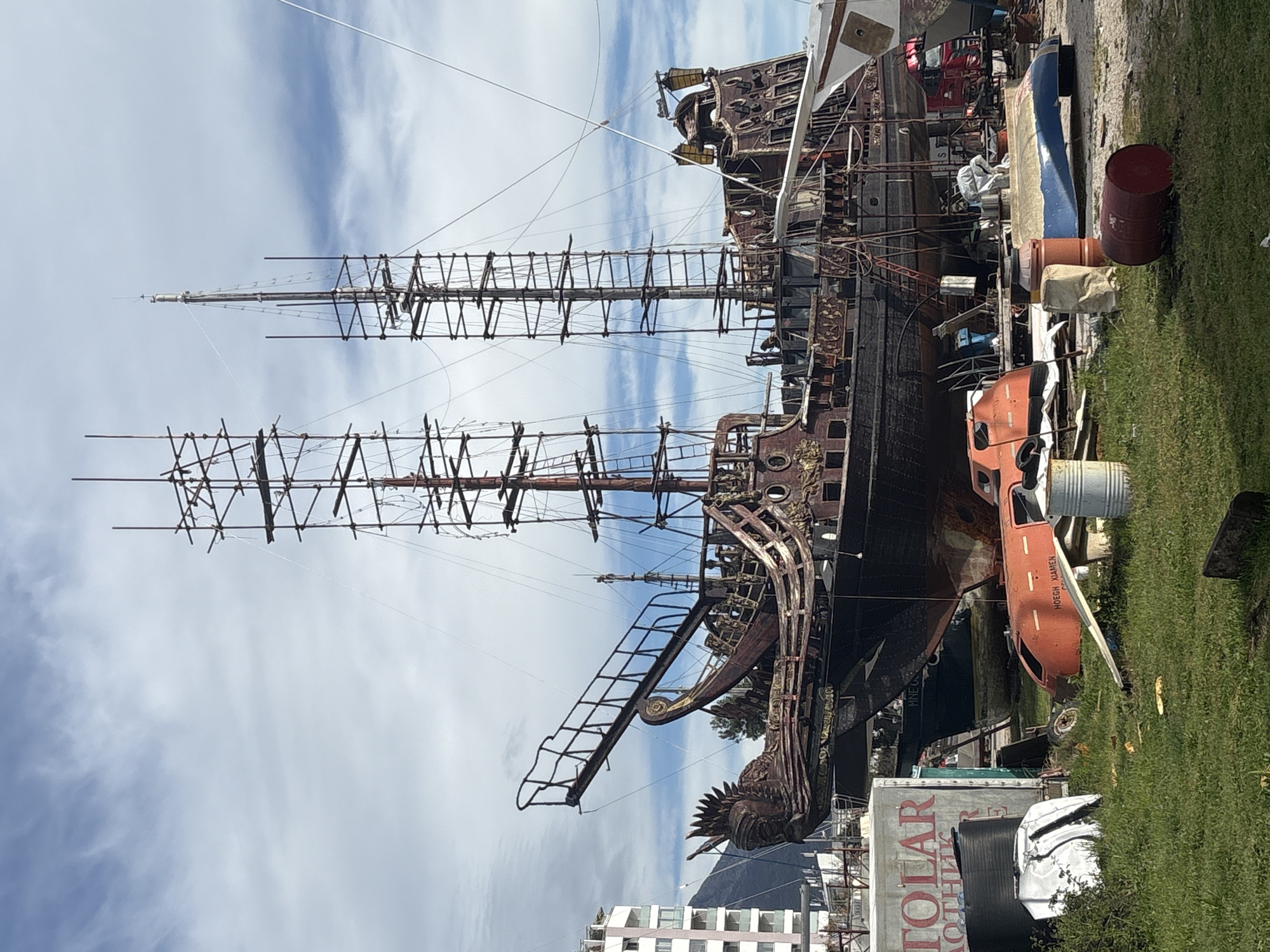 The image shows a large, disused sailing ship in a state of disrepair. The ship's hull is heavily rusted, and its rigging is incomplete. The ship is surrounded by various pieces of junk and debris. A bright orange lifeboat and red drum are visible alongside the ship. In the background, there are buildings and the sky. The ship is located in the city of Bar, Montenegro. The sign "TOLAR ROTHLE" is visible in the lower left corner.