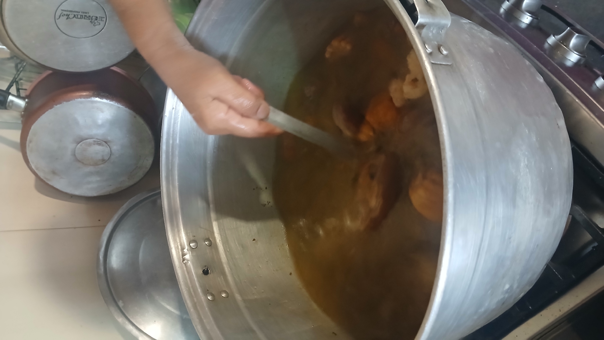 A hand is stirring the contents of a large, cylindrical, silver metal pot, possibly aluminum, filled with a dark broth containing pieces of meat. The pot is situated on a gas stovetop. To the left of the large pot are two smaller pots, one silver with the word "Eurochef" visible and the other copper-colored, stacked on top of each other. The scene appears to be in a kitchen. The city of Altagracia, Venezuela is mentioned, but there are no identifiable elements in the image to confirm its location.