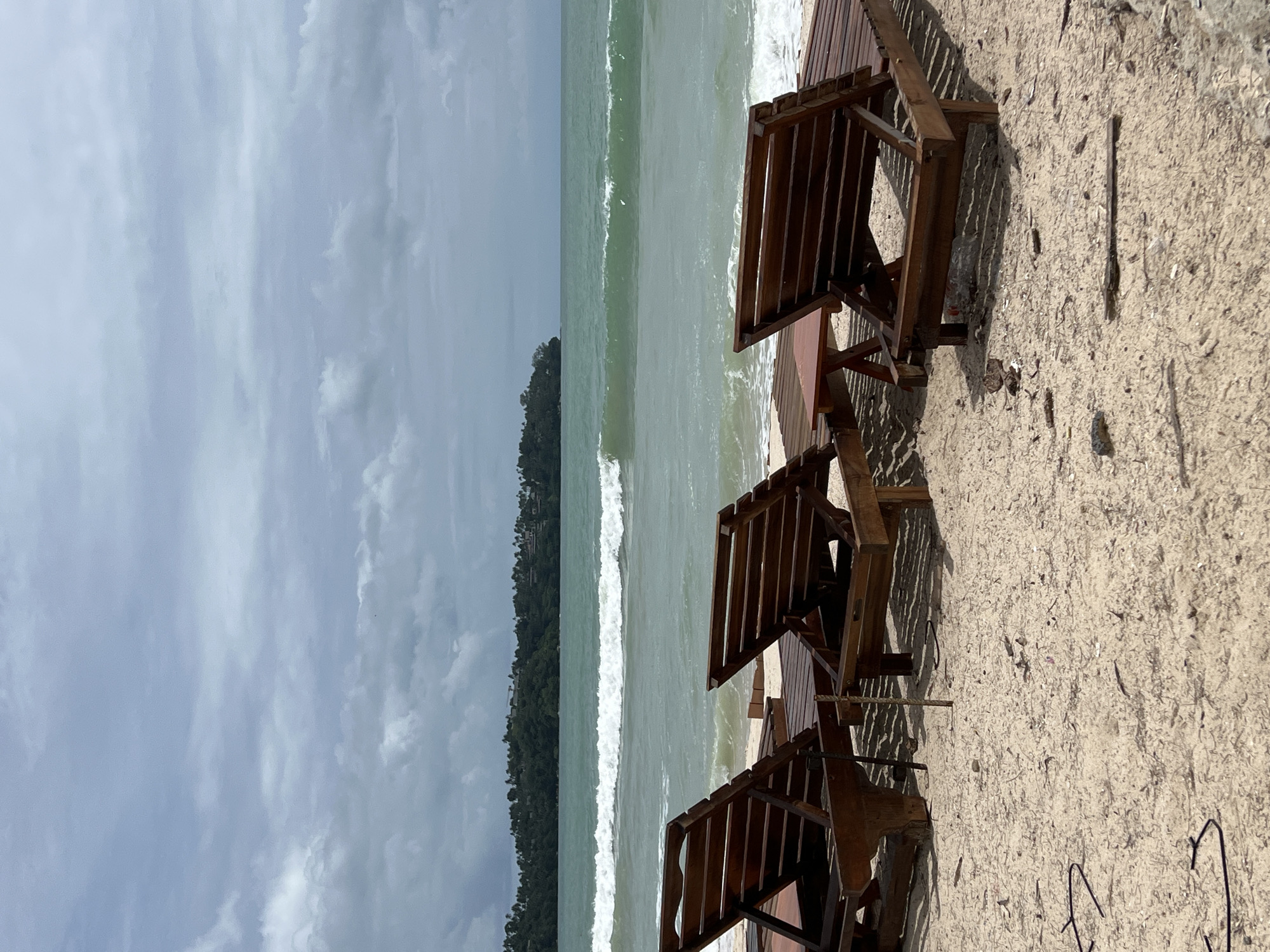 The image shows three empty wooden beach chairs arranged on a sandy beach next to a wooden deck. The chairs face the ocean, where a small, tree-covered island is visible in the distance. The sky is overcast with grey clouds, and the water appears light green. The location appears to be Unknown, Unknown.