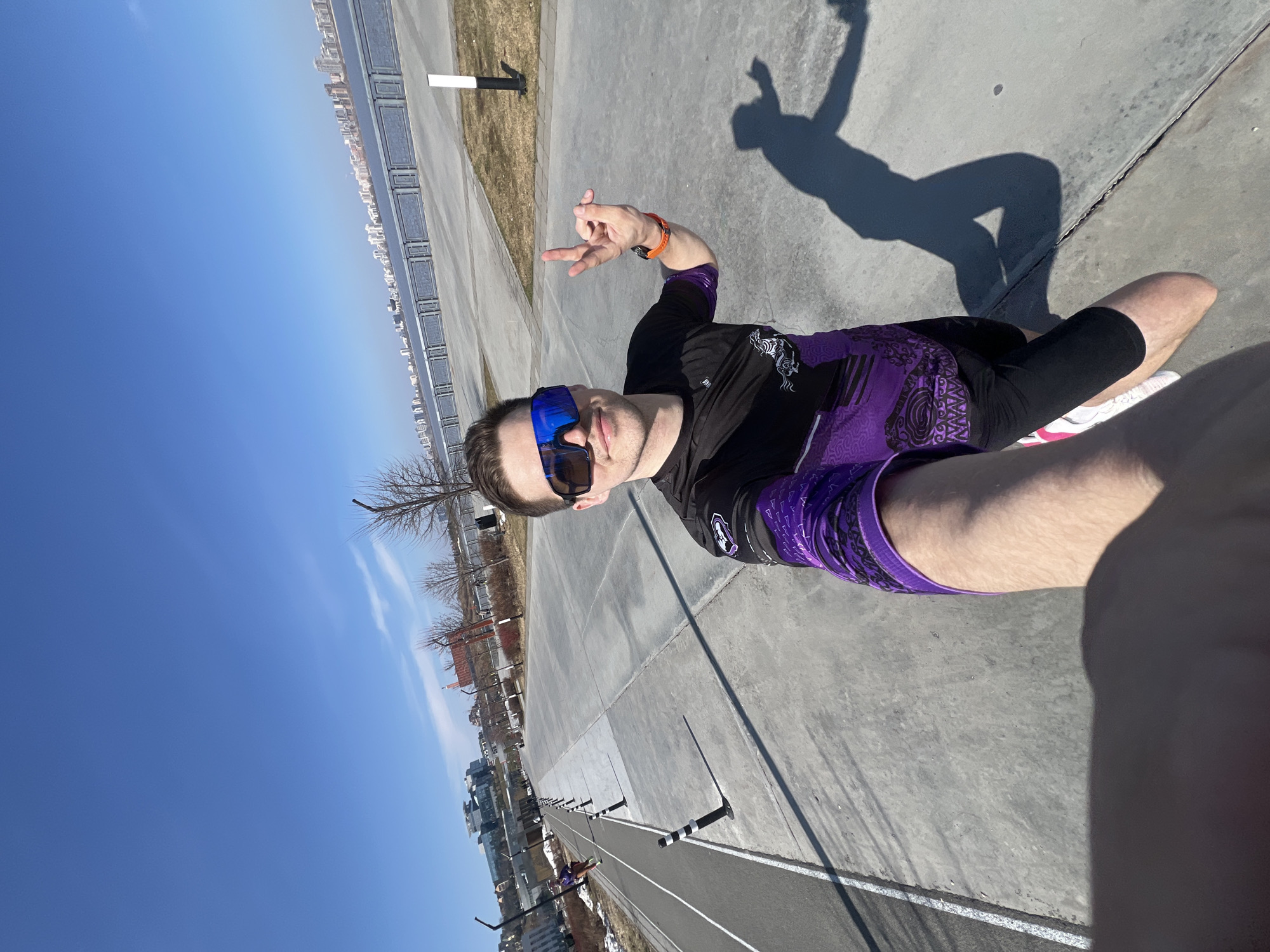 The image is a selfie taken by a person outdoors, likely on a sunny day, with a blue sky visible overhead. The person is wearing a black and purple athletic top, dark athletic shorts, and blue mirrored sunglasses. An orange watch is visible on their wrist. They are making a hand gesture.
The person appears to be on a concrete surface alongside a running track, indicated by white lines and black and white poles. In the background, a city skyline is visible, with a row of tall buildings.
Based on visual characteristics, the location is likely to be Kazan, Russia, specifically along the Kazanka River embankment. The background buildings are consistent with the cityscape of Kazan.