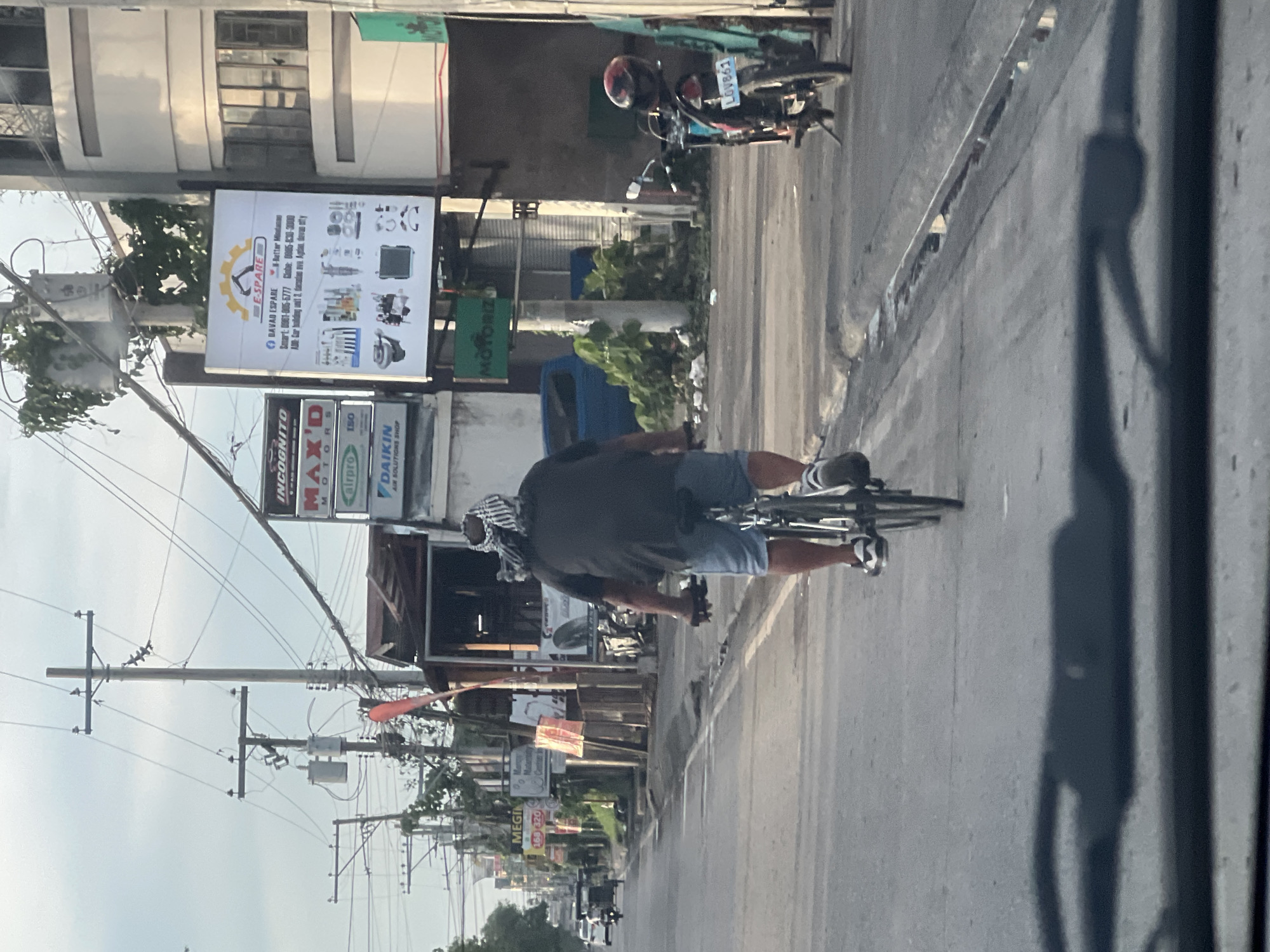 A man wearing a gray shirt, shorts, shoes, and a head covering is riding a bicycle on a gray asphalt road. The scene appears to be in Davao City, Philippines, indicated by the presence of local signage and businesses. Visible signage includes "E-SPARE," "MOTORIZ," "DAIKIN," "INCOGNITO" and "MEGIN". A motorcycle is parked along the roadside. Utility poles and overhead wires are visible. The shadow of the photographer or camera is present in the foreground.