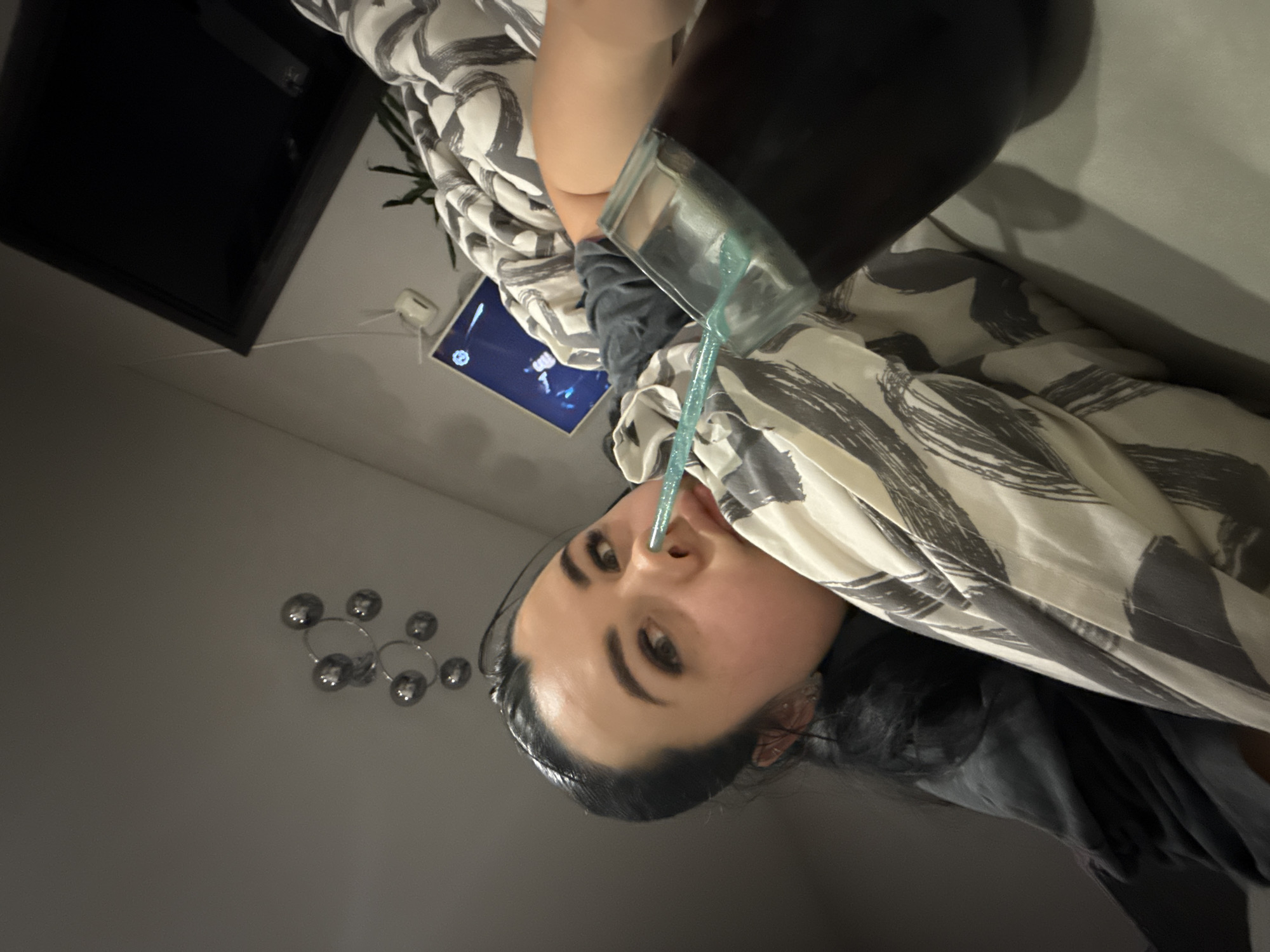 A woman is lying on a bed with a patterned comforter, holding a glass with a blue straw in her mouth. The woman has dark hair pulled back and is wearing a gray shirt. To her left is a wall with a silver circular light fixture. Further in the background, a television is mounted on the wall. The location is Chaikovsky, Russia.