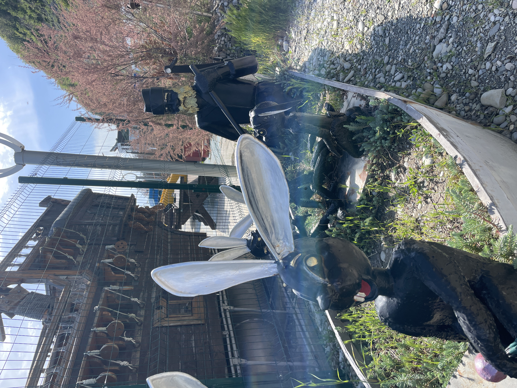 The image shows an outdoor scene in Olginka, Russia, featuring whimsical sculptures and objects within a fenced area. The primary focus is on two dark-colored sculptures with large, white, metallic wings, resembling insects or fantastical creatures. The sculptures appear to be made of metal or other durable material and are positioned among green foliage and a gravelly surface. Behind the sculptures, a chain-link fence is visible, partially obscuring a multi-level wooden structure or ship. The background includes glimpses of other objects and greenery. The environment appears to be a garden or decorative space. The sky is visible in the upper-left corner, suggesting a sunny day.