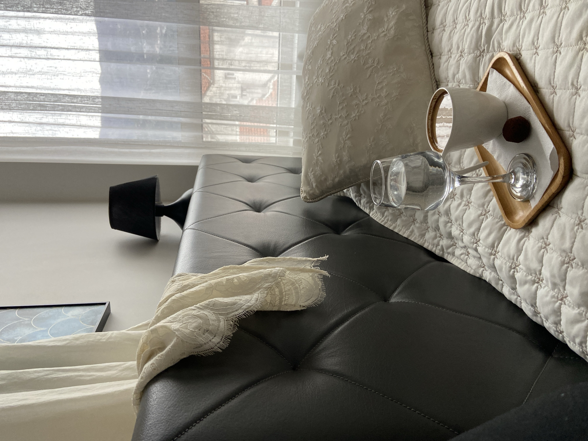 The image shows a room setting with a focus on domestic items. A black tufted sofa or ottoman is prominently featured. On the right side of the sofa is a white quilted blanket and a decorative pillow. On top of the blanket, there is a wooden tray holding a white mug, a dark round item (possibly a truffle), and an overturned wine glass. A lace scarf drapes across the sofa. In the background, a window with sheer curtains provides a view of what appears to be buildings. A small black lamp is also visible on the left side of the sofa. The specific location in Budva, Montenegro, cannot be determined from the visual information provided.