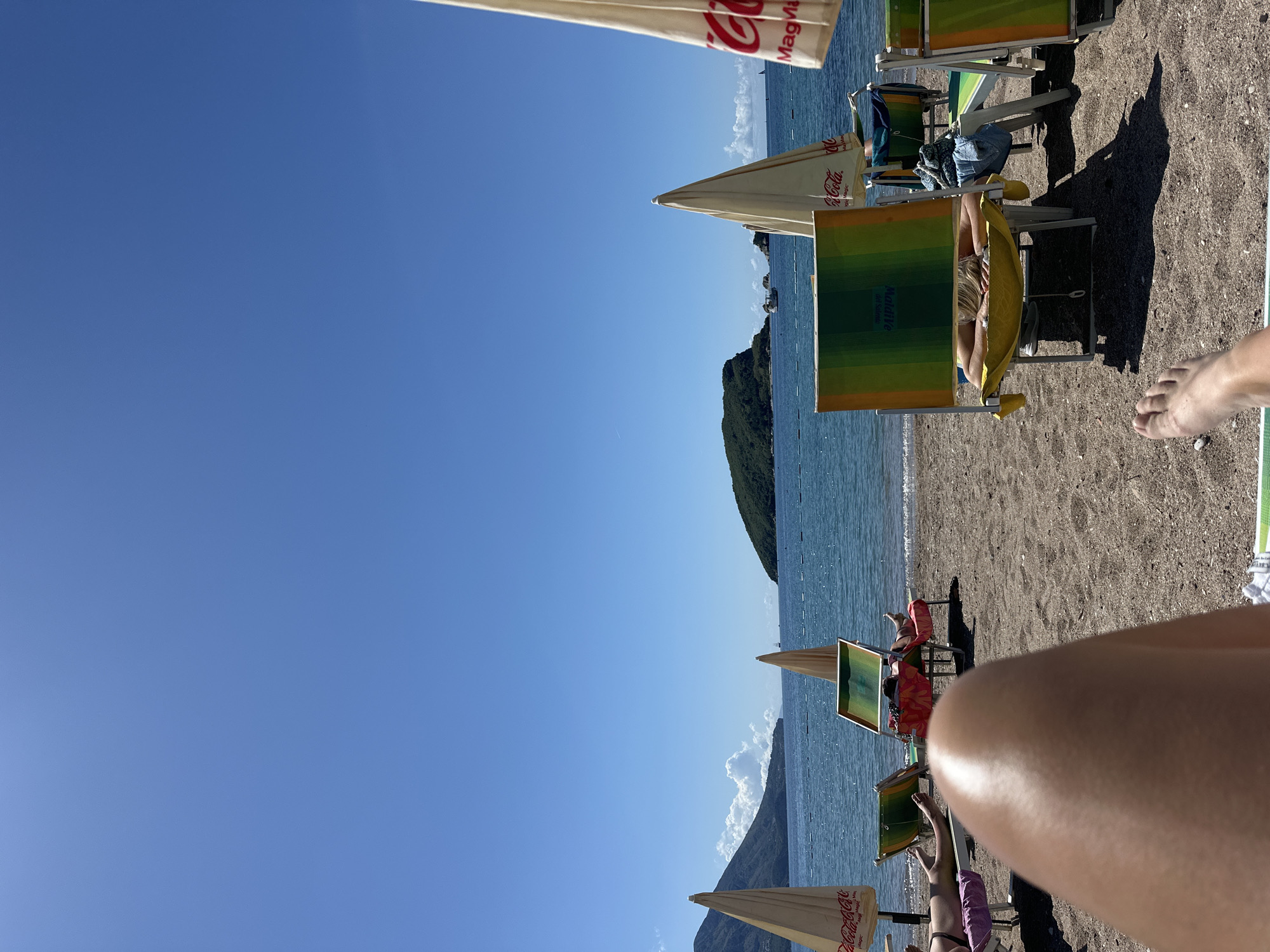 The scene is a beach in Budva, Montenegro, with the azure sea and clear blue sky dominating the upper left and center of the frame. Several beach umbrellas with Coca-Cola logos are visible along the shoreline, interspersed with colorful beach chairs. A woman is relaxing in one of the beach chairs, wearing yellow clothing. A bare foot of another person is visible in the upper right. A leg is in the foreground obscuring part of the view. A small, green-covered island is in the background, adding to the coastal scenery. The sand is coarse and dark.