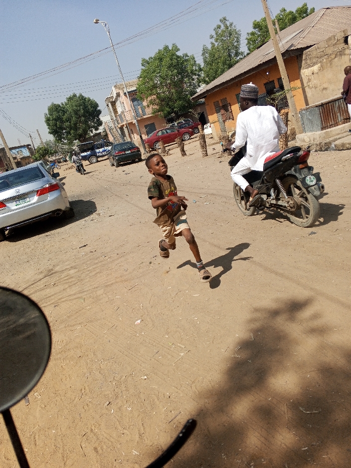 The image depicts an unpaved, dusty street scene in Gama, Nigeria. In the foreground, a male child, estimated to be 5-8 years old, is captured mid-stride, running from the right towards the left. He wears a dark t-shirt with a graphic, light-colored shorts, and sandals, displaying an intense facial expression. His shadow extends to his right. A dark, circular object, consistent with a vehicle mirror, is partially visible in the lower-left quadrant.
In the mid-ground, a male individual in a white traditional garment and white cap is riding a black motorcycle, moving from left to right, away from the child.
The background features various buildings, including a multi-story structure on the left and single-story buildings with weathered, light brown/orange walls on the right. A silver sedan (Honda Accord) is parked on the left, and further back, a blue and white police vehicle is visible, along with other parked cars (including a red sedan) and another distant motorcycle. A street light pole stands centrally. Green trees are interspersed throughout the background. The scene is under clear skies with strong sunlight, indicated by pronounced shadows. Wooden stakes are visible along the right side of the road.