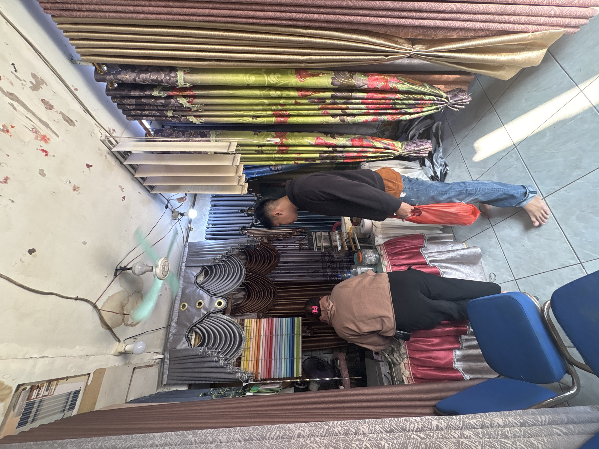 In a cluttered curtain shop in North Jakarta, Indonesia, a female customer wearing a brown hooded sweatshirt and black pants is browsing fabric samples displayed on a table covered with a red tablecloth. A male customer in a black hooded sweatshirt and blue jeans, carrying a red plastic bag, stands near the fabric display, looking at the curtains. The shop is filled with various colorful curtains, and a ceiling fan is visible. The shop appears to be an indoor space with tiled flooring and a worn ceiling.