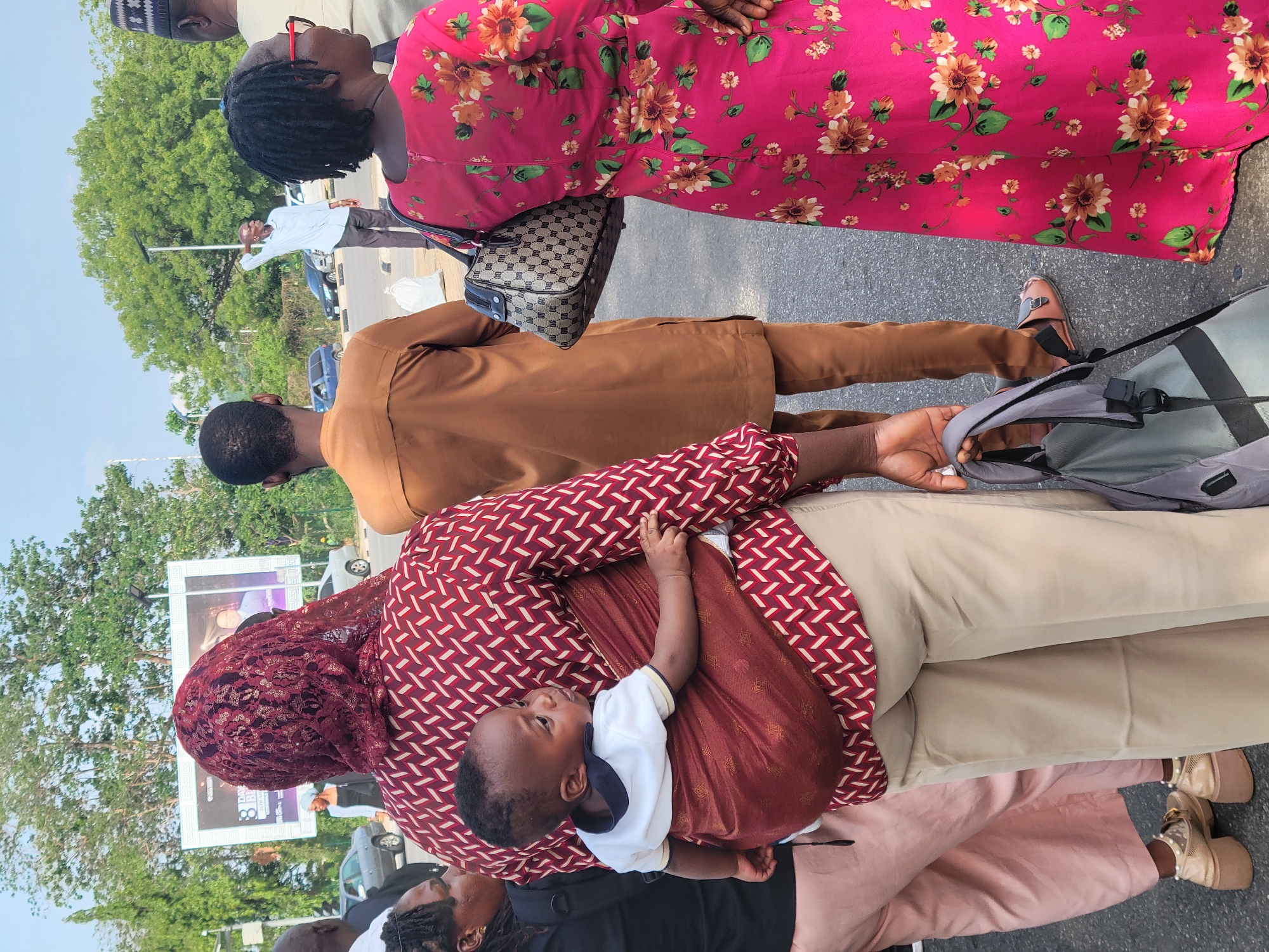 A group of people stand on a paved surface, likely a sidewalk or street, in Abuja, Nigeria. A woman in the foreground carries a baby in a sling. The woman wears a maroon and white patterned top, beige pants, a maroon lace head covering, and tan platform shoes. The baby wears a white shirt and a maroon sling. Beside her stands a person in a brown shirt and sandals. Next to them is a woman wearing a pink floral dress and carrying a patterned handbag. Behind them are other people, a green grassy area, trees, streetlights, and a large billboard. Cars are visible on the street. The sky is bright and clear.