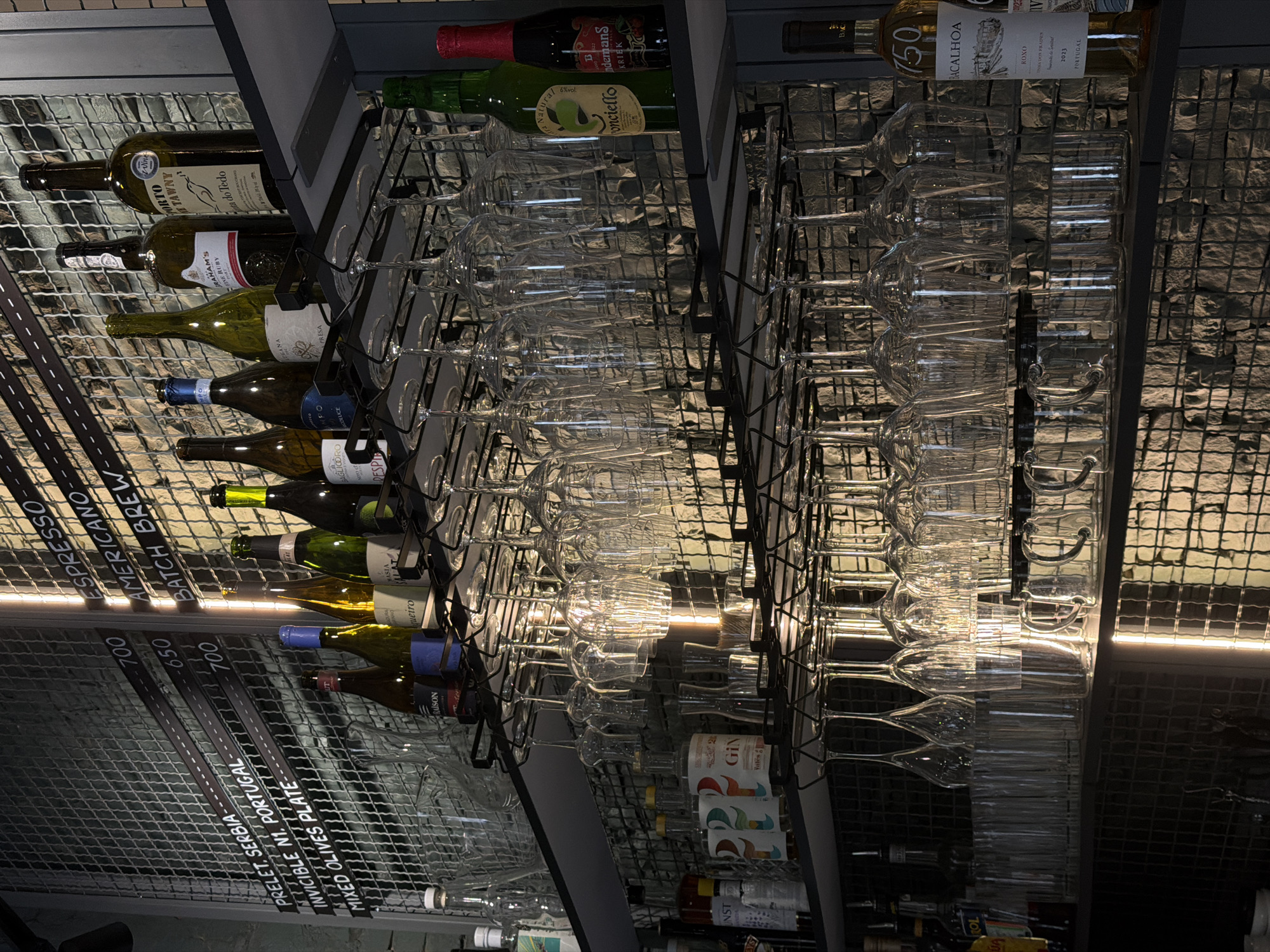 The image depicts the interior of a bar or restaurant, likely located in Belgrade, Serbia, as suggested by the menu items visible on the wall. The focus is on shelves stocked with bottles and glassware. On the top shelf, several bottles of wine and spirits are arranged, including a bottle of "Porto Tawny" and a "Graham's Fine Ruby." A menu board behind the bottles lists items like "PRELET, SERBIA," "INVISIBLE N1, PORTUGAL," and "MIXED OLIVES PLATE," along with prices.
Below the bottles, a rack holds numerous wine glasses suspended upside down. The second shelf down displays more bottles, including various types of gin, one labeled "GIN" with colorful graphics, and two bottles of what appear to be alcoholic beverages, one green with a label featuring an apple and "Non-alcoholic," and another red bottle labeled "Kriek."
The third shelf features another set of suspended glassware, and the shelf below that holds a collection of clear glasses, some appearing to be tumblers and others smaller shot glasses. On the right side of the second shelf from the top, a bottle labeled "750 BACALHOA ROXO" is visible, with "2023 PORTUGAL" printed on its label. The background consists of a stone-tiled wall with a wire grid overlay. The lighting is dim, with some artificial light sources casting a warm glow. There are no people visible in the frame, and the scene is static, implying a captured moment in time.
