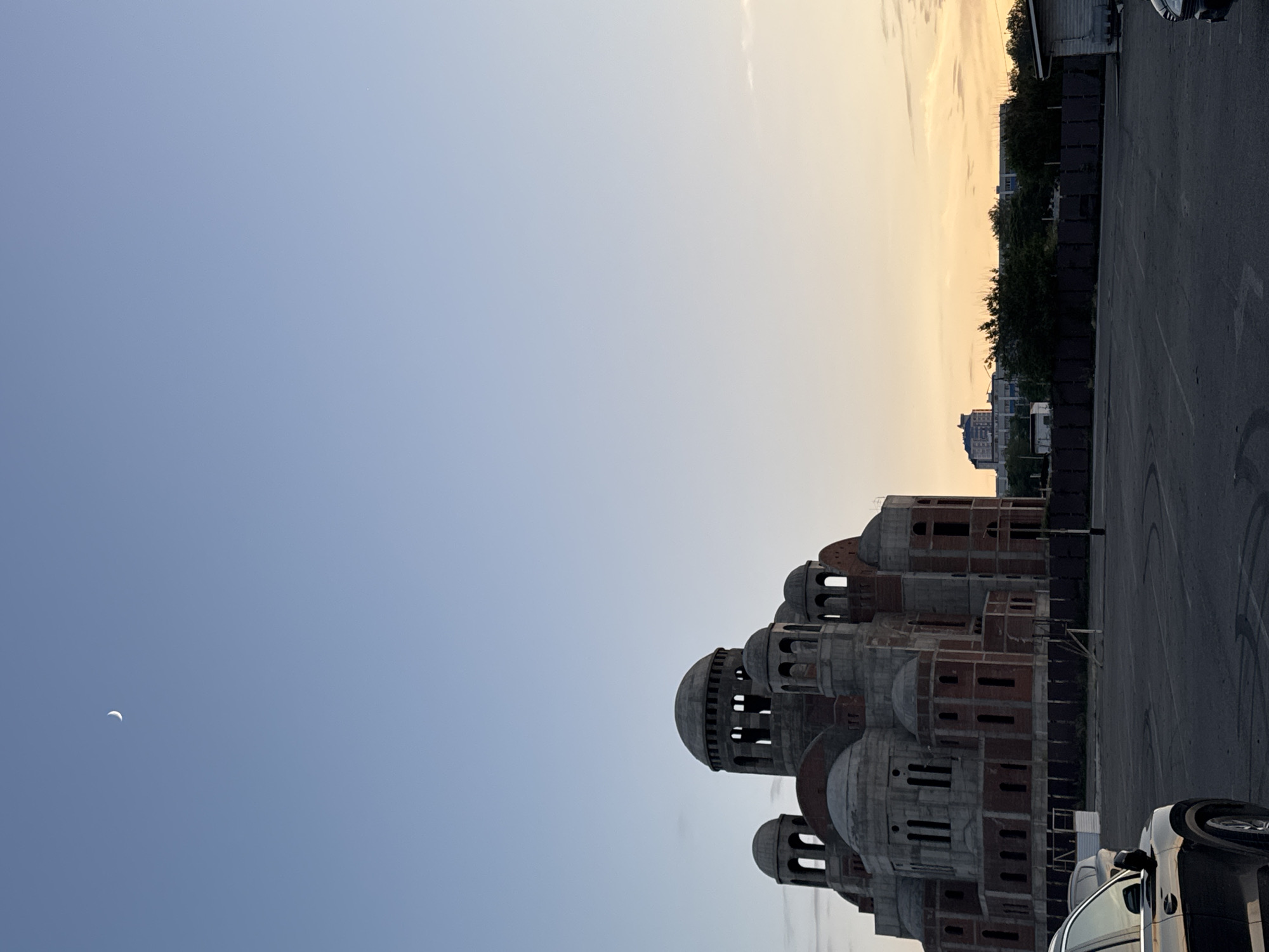 This media file depicts a partially constructed religious building, possibly a church or cathedral, with multiple domes and intricate architectural features. The building's exterior is a mix of exposed brickwork and concrete, indicating ongoing construction. The scene is set against a twilight sky with a soft gradient of blue and yellow, suggesting either dawn or dusk. A crescent moon is faintly visible in the upper left portion of the sky.
To the right of the building is an asphalt parking lot, marked with white lines. A few vehicles are visible in the parking lot, including the rear of a white car in the foreground. Behind the parking lot, a low, dark fence runs horizontally, and beyond that, a line of trees and some distant buildings can be seen. The overall atmosphere is calm and serene, with the construction site appearing quiet.
The location is identified as Novorossiysk, Russia. The time of day, indicated by the sky, is likely early morning or late evening. No people are clearly visible in the frame. The image is in portrait orientation.