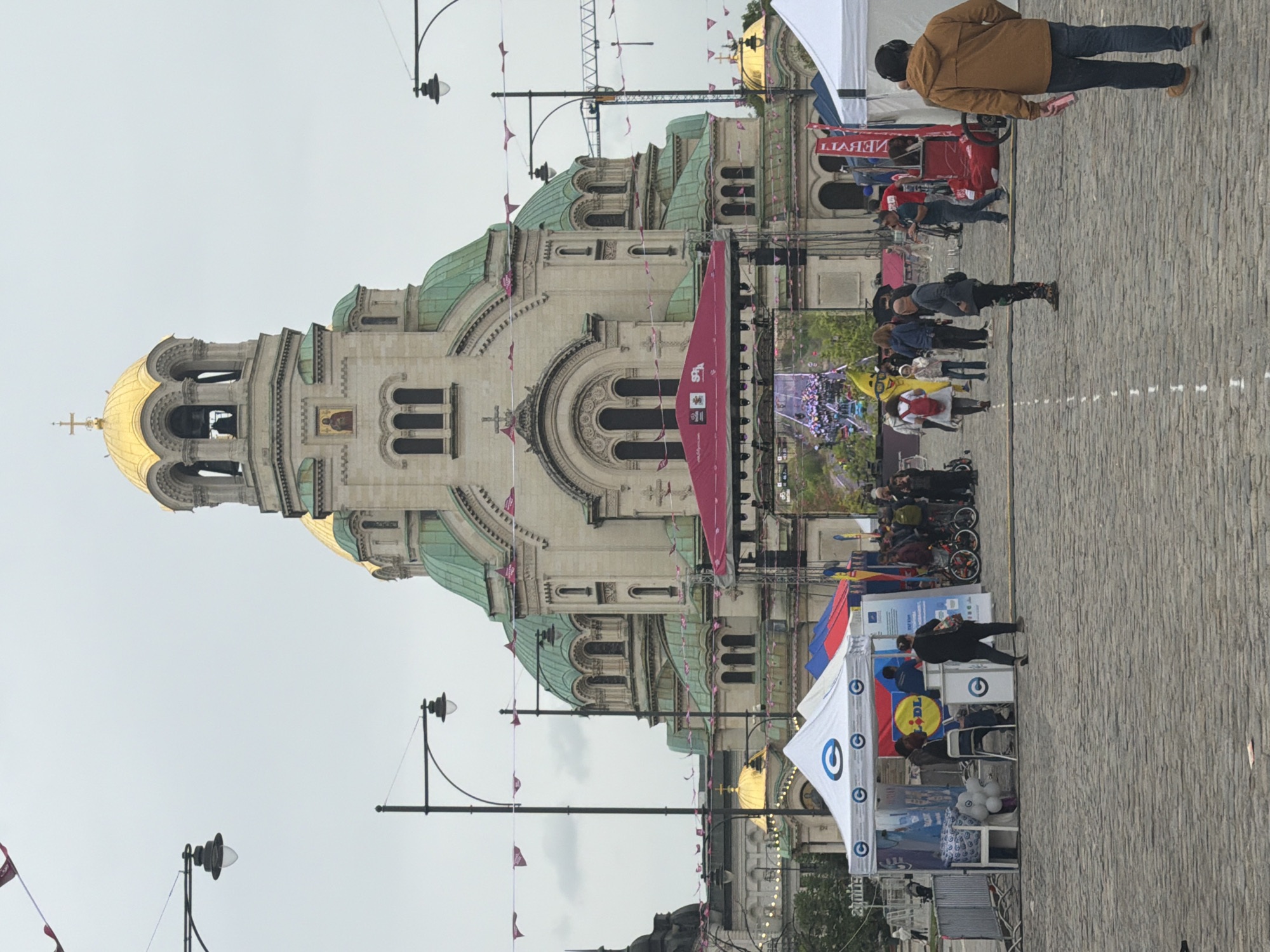 The image captures the Alexander Nevsky Cathedral in Sofia, Bulgaria. The cathedral's domes and detailed facade are prominently displayed. A crowd of people is present in front of the cathedral, amidst event tents and displays. The ground is paved with cobblestones. Banners and flags adorn the area. People are walking and interacting with the event setups.