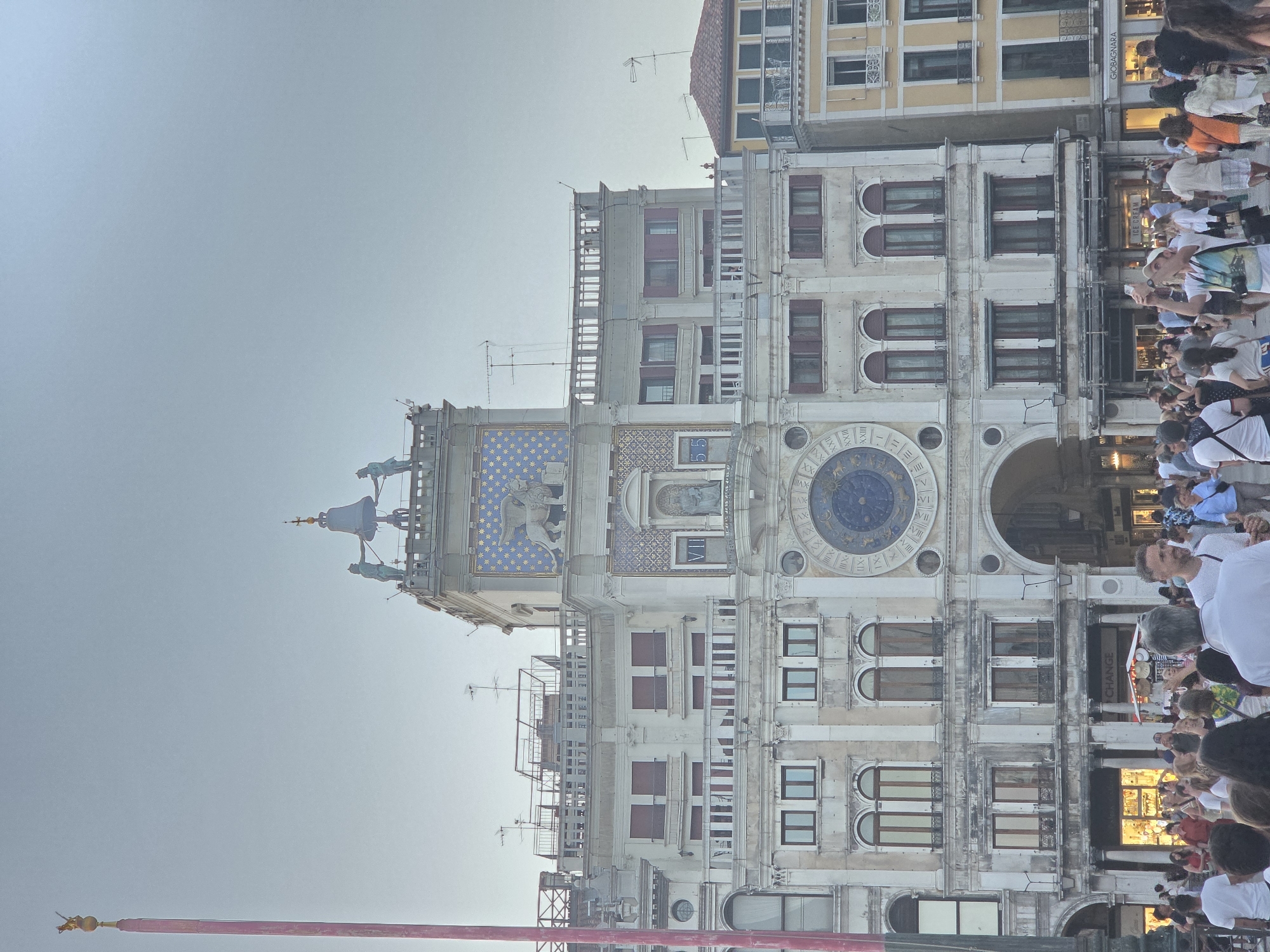 The image shows the Torre dell'Orologio (Clock Tower) in St. Mark's Square (Piazza San Marco) in Venice, Italy. The tower is a prominent architectural landmark, featuring a large astronomical clock face and statues on its upper level. A crowd of people is gathered in the square below, some looking at the tower, others engaged in conversation or taking pictures. The scene appears to be set during the daytime, with a clear sky. The architecture of the surrounding buildings and the presence of a canal or waterway are not clearly visible in this particular shot, but the overall context suggests a bustling tourist area in Venice.