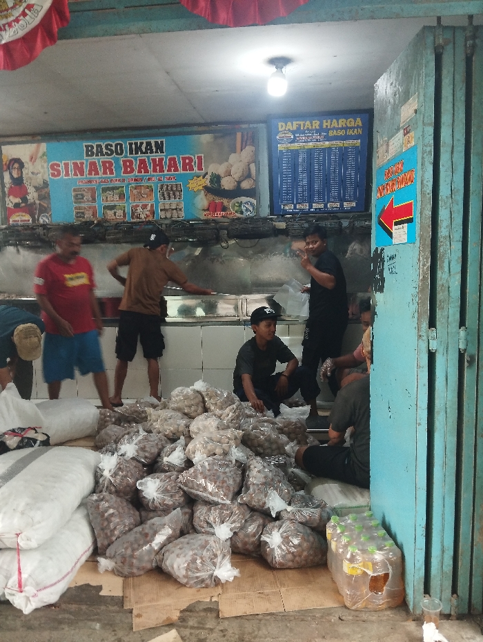 This image captures a bustling indoor commercial space in Baleendah, Indonesia, likely a production or distribution point for meatballs. The scene is dominated by several men, appearing to be workers, engaged in handling large quantities of food.
In the foreground and mid-ground, numerous clear plastic bags filled with meatballs (baso) are piled high on cardboard sheets on the floor, alongside several large white woven sacks, possibly containing more meatballs or raw ingredients. A few men are squatting or standing around these piles, suggesting they are sorting, packing, or moving the products. In the background, three men stand behind a counter or work surface. One man on the right, wearing a black shirt, looks towards the camera and makes a hand gesture.
Overhead, a bright fluorescent light illuminates the scene, indicating an indoor setting during what appears to be daytime. The walls in the background feature tiled surfaces, common in food preparation areas. To the right, a folded blue metal accordion-style door or shutter marks the entrance or exit.
Visible text includes a large banner across the back wall that reads "BASO IKAN SINAR BAHARI" (Fish Meatballs Sinar Bahari), featuring images of various products. Next to this, a blue sign displays a "DAFTAR HARGA BASO IKAN" (Price List Fish Meatballs). A smaller, bright blue sign with a red arrow pointing left is affixed to the blue shutter, with some unreadable Indonesian text. The overall impression is that of a local, high-volume operation focused on the production and sale of fish meatballs.