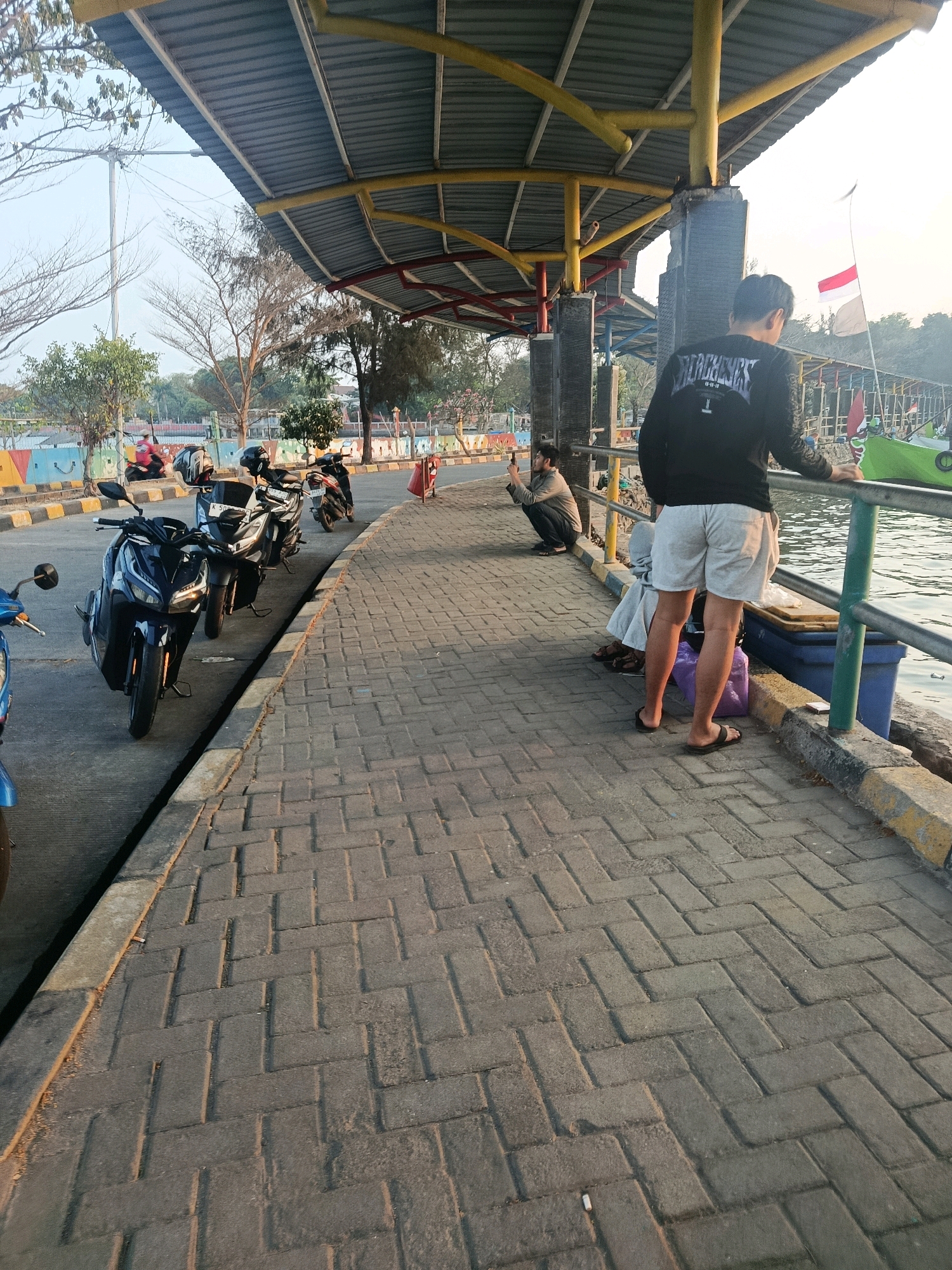 The image depicts a waterfront promenade in Jepara, Indonesia, with a covered walkway and a paved path. In the foreground, several motorcycles are parked along the edge of the walkway. A person in a black long-sleeved shirt and light-colored shorts stands near the railing, looking out at the water. Another person is seated on the ground nearby, taking a photograph with a smartphone. In the background, more people are visible, along with a boat and a flagpole with an Indonesian flag. The scene appears to be set during the late afternoon, with soft sunlight casting long shadows.