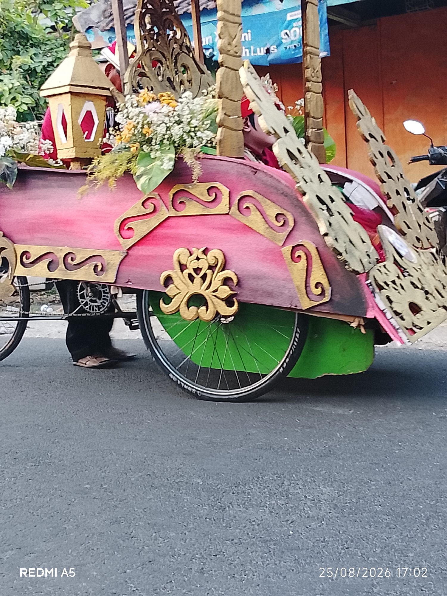 This is a close-up, low-angle shot of a decorated bicycle rickshaw, likely part of a parade or cultural event in Semanding, Indonesia. The rickshaw is adorned with vibrant pink and gold paint, featuring ornate scrollwork and floral arrangements. A gold lantern with white hexagonal accents is visible on the front, along with lush greenery and white flowers. The back of the rickshaw has intricate, carved wooden elements that resemble wings or flames, painted in gold. A person's legs and feet, wearing sandals and dark pants, are visible, suggesting they are operating or sitting in the rickshaw. The vehicle's large green wheel with a black tire and silver spokes is prominent in the foreground. The background shows a blurred orange wall, possibly a building, and hints of greenery. A motorcycle is partially visible on the right side. The date and time stamp at the bottom right indicates "25/08/2026 17:02," and "REDMI A5" is watermarked at the bottom left. The scene is set on an asphalt road.