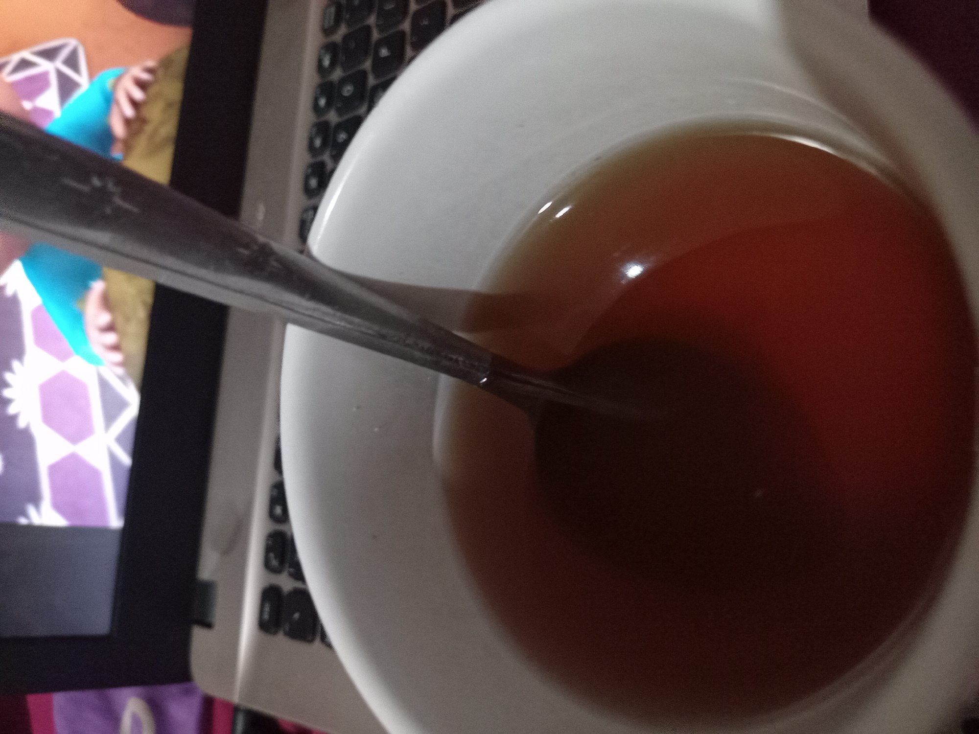 The image shows a close-up of a white bowl filled with a dark amber liquid, possibly tea. A metal spoon is submerged in the liquid, angled towards the right side of the frame. The bowl rests on a silver laptop with visible keys. In the background, the laptop screen displays a colorful, geometric pattern with figures that appear to be cartoon characters. The overall setting suggests a casual indoor environment, possibly a home or dormitory room. The image does not provide any specific geographical information that would allow for pinpointing a location in Claket, Indonesia.