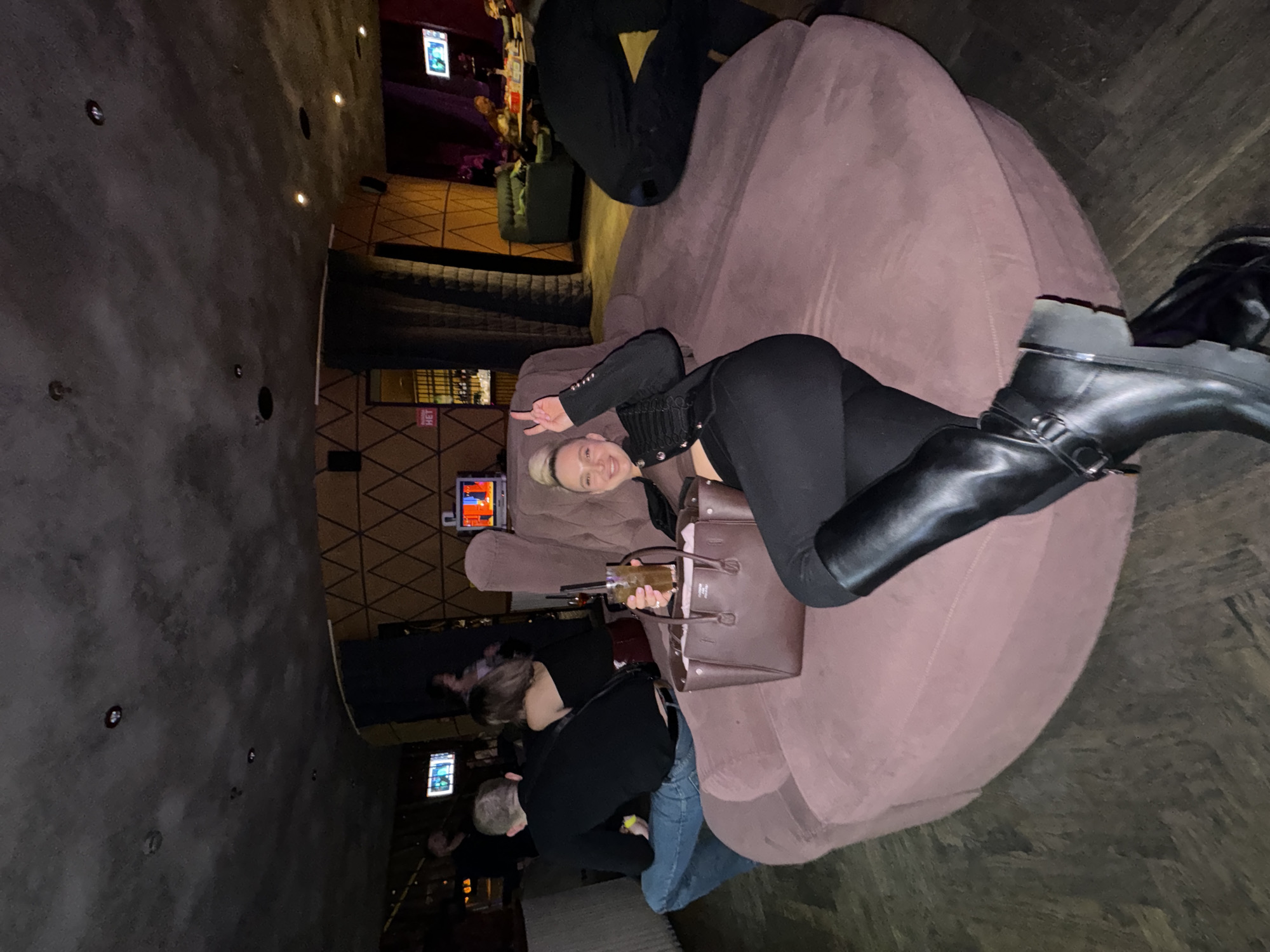 A photo taken in what appears to be a bar or lounge in Moscow, Russia, shows a woman lying on a large, plush, round couch. She is wearing black leggings, a black jacket with decorative silver details, and black boots. She is holding a brown handbag and a glass with a drink. Her left hand is raised, displaying a peace sign. Two other people are partially visible, one kneeling on the floor and another sitting on the couch. There are television screens in the background, typical for entertainment venues. The interior design features a gray textured wall with embedded lights and wooden panels.