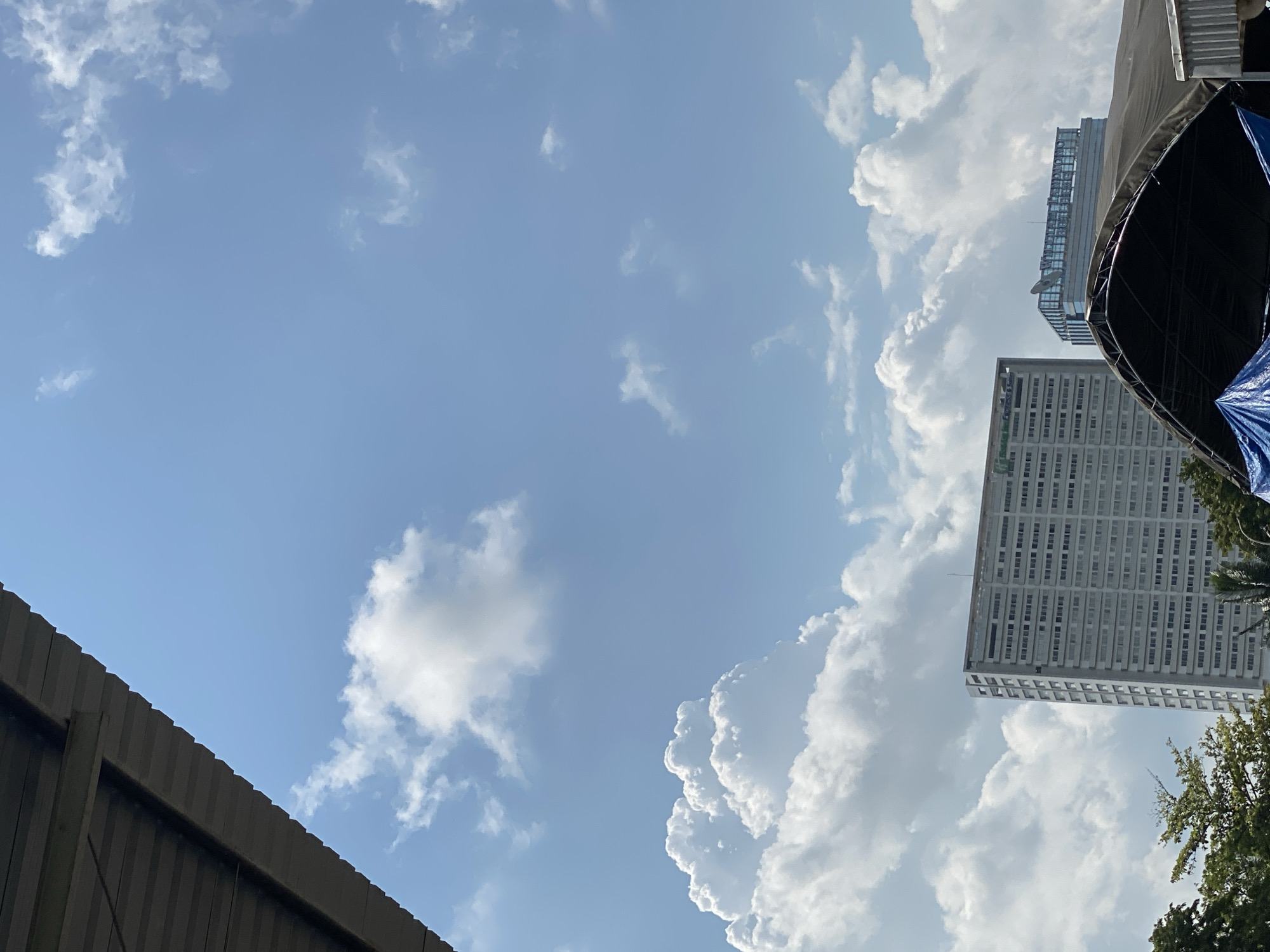 The image captures a daytime scene in Gambut Barat, Indonesia, dominated by a bright blue sky filled with fluffy white clouds. In the foreground, a portion of a grey, corrugated metal roof overhangs the frame from the top left, casting a shadow. Below and to the right, two modern buildings rise against the sky. The taller building on the left is a light-colored, multi-story structure with numerous windows, appearing to be an office building or hotel. A sign with green text is visible near its top. To the right of this building is a shorter, glass-fronted building with a prominent rooftop sign. In the bottom portion of the image, a large, dark, curved canopy or tent structure partially obscures the view of trees and other objects below, with a blue tarp visible underneath it. The overall impression is an urban landscape under a clear, sunny sky with scattered clouds.