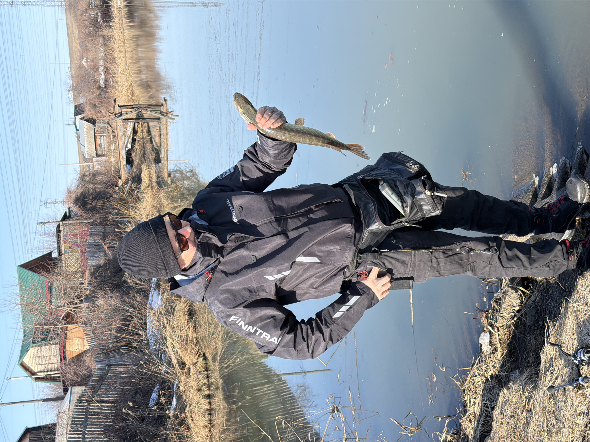 A man is holding a fish in his right hand. He is wearing a black beanie, sunglasses, a black jacket with "Finntrail" written on the left sleeve, and black pants. He is standing on a bank next to a body of water. In the background are buildings and dry grass. A fishing rod is on the ground next to him. The location is near Chaikovsky, Russia.