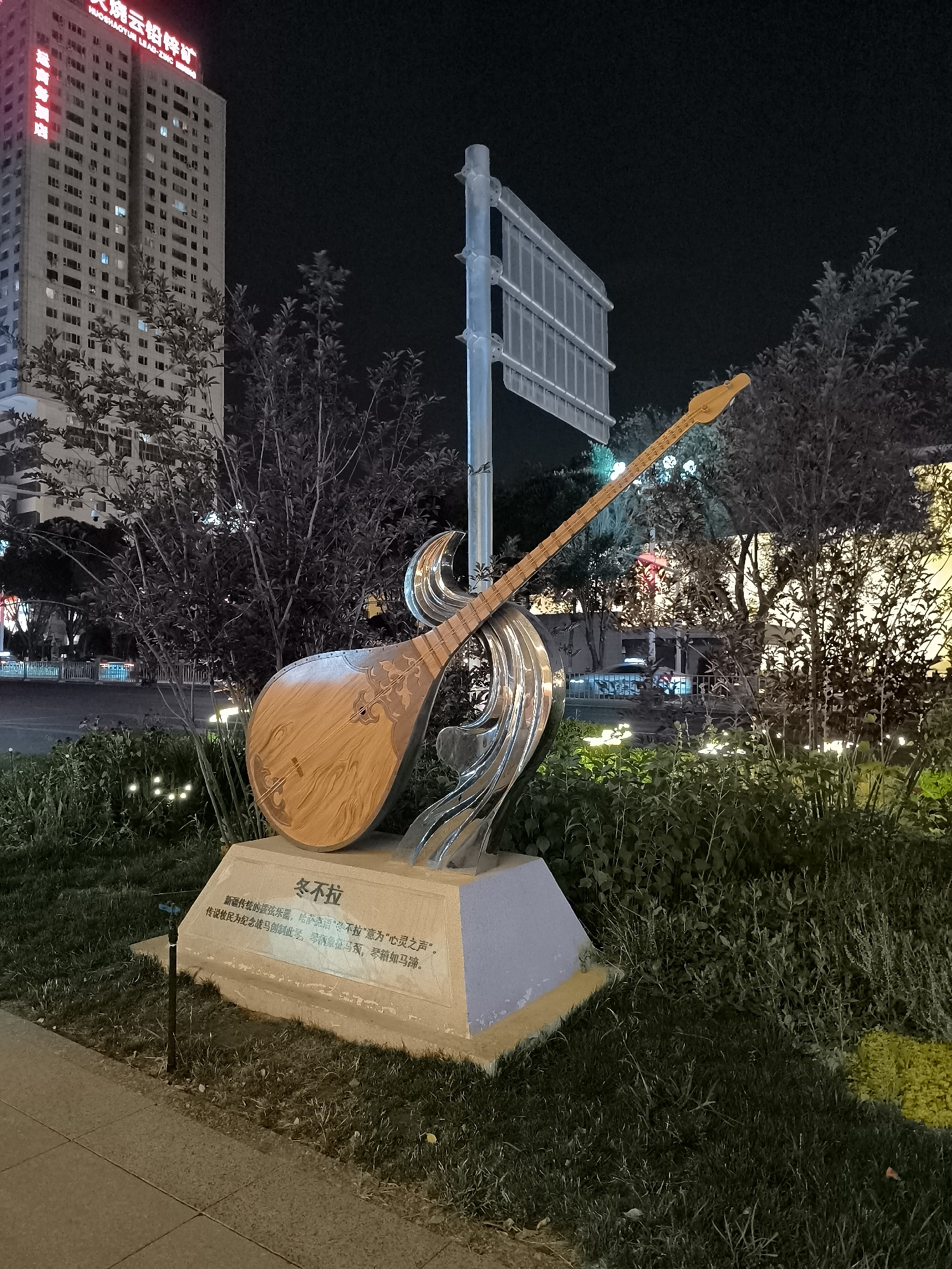 The image shows a sculpture of a traditional Kazakh instrument, the "Dombra", on a pedestal, located in front of a tall building in Urumqi, China. The sculpture is made of wood and metal, and it is illuminated by streetlights at night. There is text on the pedestal, which appears to be in Chinese and possibly Kazakh. The surrounding area is landscaped with grass and bushes, and there are trees in the background. A street with cars is visible in the distance, and a street sign is mounted on a pole next to the sculpture. The building in the background has a red neon sign at the top that reads "Huo Shaotun Lead-Zinc Mine." The sculpture is a public art installation, likely intended to represent the local culture and heritage of Urumqi, which has a significant Kazakh population.