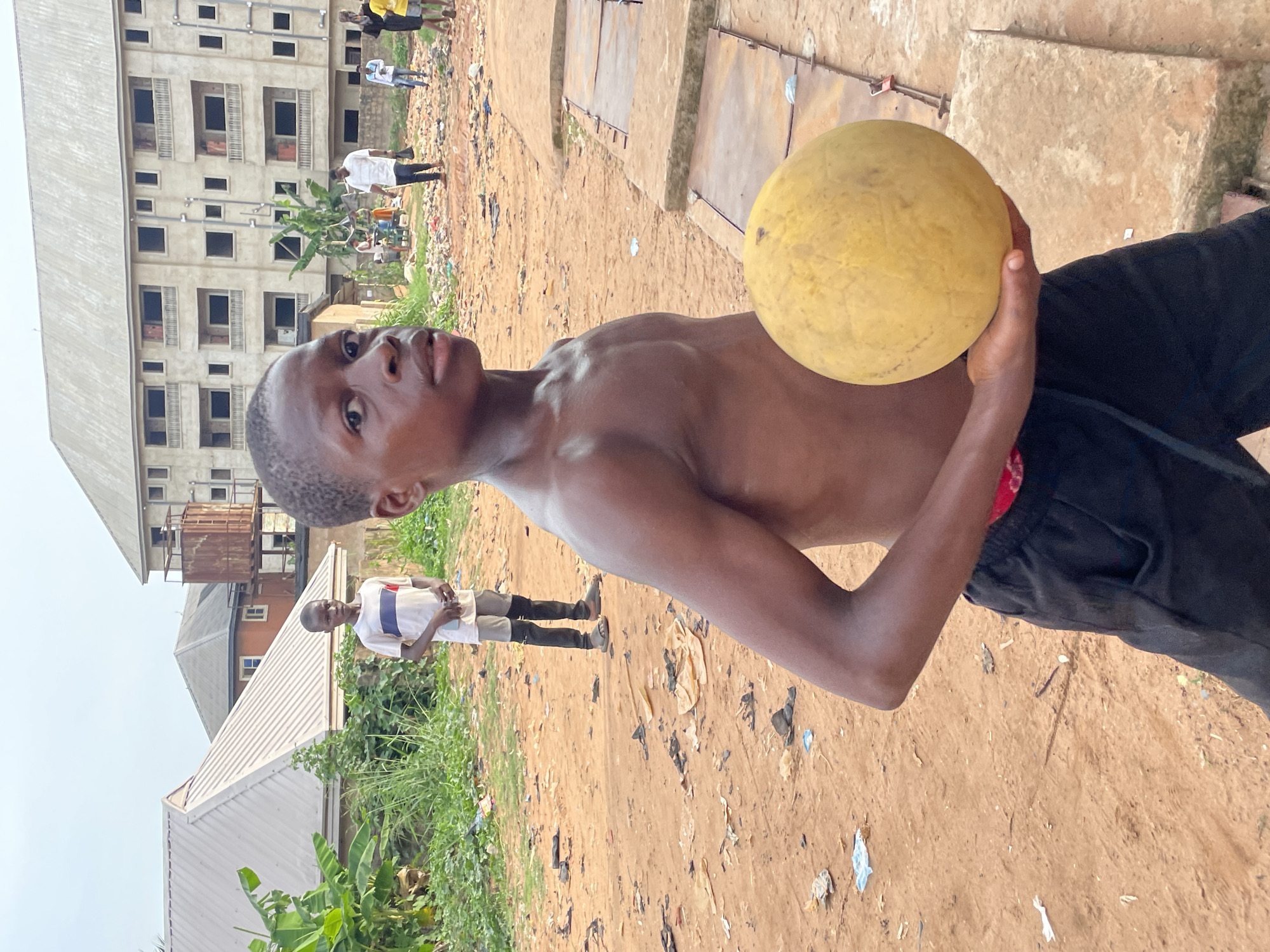 The image depicts an outdoor scene in what appears to be an urban or semi-urban area, consistent with Onitsha, Nigeria. The primary subject is a young, dark-skinned male, shirtless and wearing black trousers with a red band or fabric visible at his waist. He is positioned in the foreground, holding a large, spherical yellow fruit, resembling a melon, in his hands. He looks directly at the camera with a neutral expression.
The background features reddish-brown, dry earth, sparsely covered with green vegetation, including banana plants on the left. Scattered litter and debris are visible on the ground. Several unfinished or partially constructed concrete buildings with multiple stories and open window frames dominate the left side of the background. Another building with a metal roof and a rusty water tank is visible further back. Concrete slabs or makeshift pathways are also present on the right side of the mid-ground.
In the mid-ground, other people are visible: one person in a white t-shirt and dark pants stands to the left of the main subject, and another in a striped shirt with dark knee-high coverings is further in the distance. The scene is brightly lit by natural sunlight, suggesting it is daytime with clear weather. No visible text is present in the image.