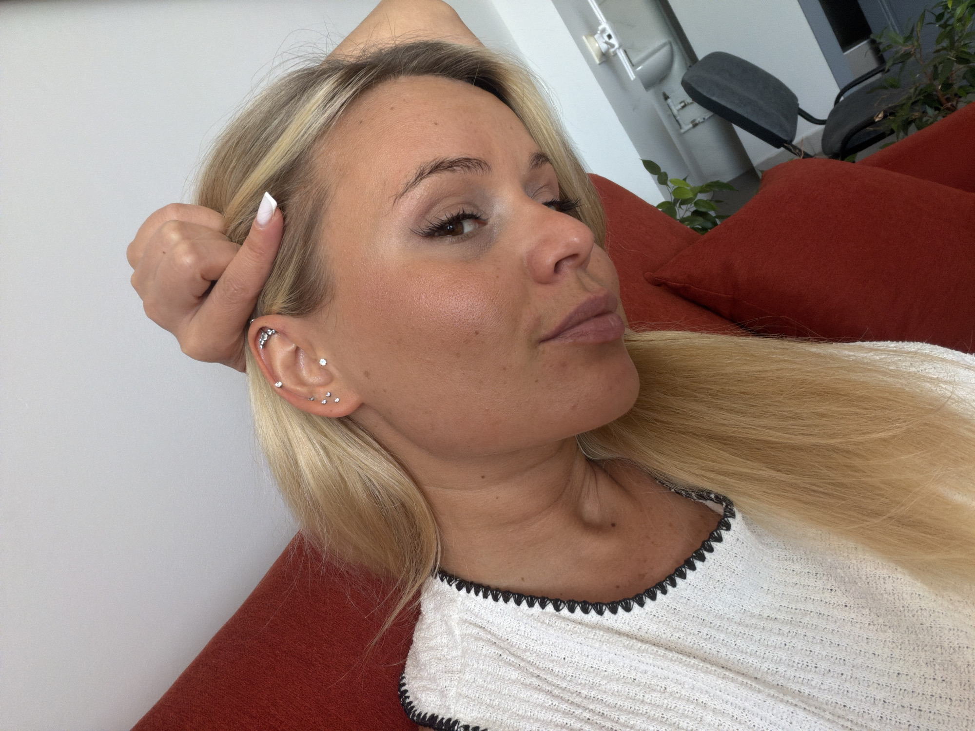 An outdoor, eye-level, close-up selfie of a woman with blonde hair. She is lying down on a red sofa, with her right arm bent and her hand holding her head. She is wearing a white top with a black trim. Her left ear is adorned with three small sparkly earrings. The background is out of focus, but shows a wall and some furniture. The image was taken in Sarapul, Russia.