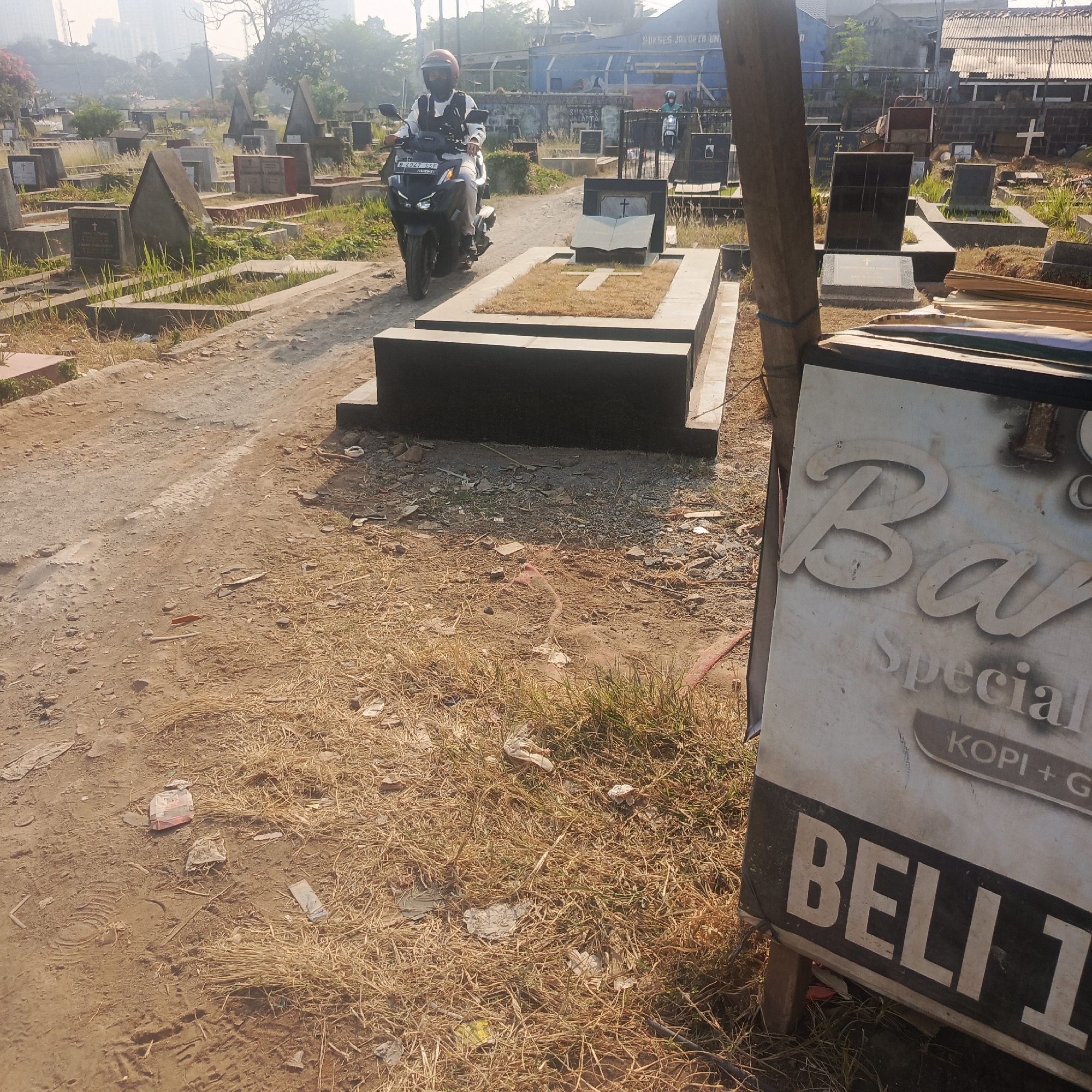 A man rides a motorcycle through a cemetery in South Jakarta, Indonesia. The cemetery is filled with gravestones of various sizes and materials, some with upright markers and others with flat tombs. Dry grass and dirt cover the ground, with scattered debris. In the foreground, a large sign displays text in what appears to be Indonesian, with the word "BELI" (buy) prominently visible. A wooden post supports the sign. The overall scene depicts a somber environment with signs of recent activity.