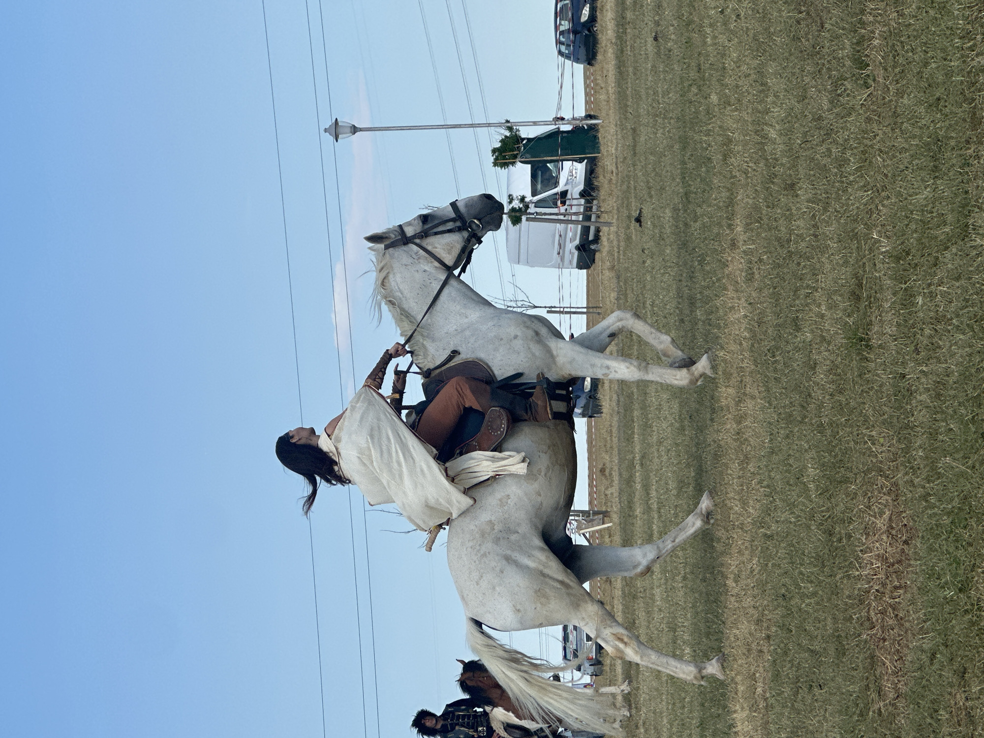 The image depicts a sunny outdoor scene in Neofit Rilski, Bulgaria, with a clear blue sky. In the foreground, a person dressed in historical attire, featuring a white cape and brown leggings, rides a white horse at a trot across a grassy field. The horse is wearing a bridle and saddle. To the left, another person on a brown and white horse, wearing a dark, ornate jacket, is partially visible. In the background, a white van, a dark car, and a lamppost are present. The ground is covered with dry grass and some darker patches, suggesting a dry spell. The overall atmosphere is that of an outdoor event or reenactment.