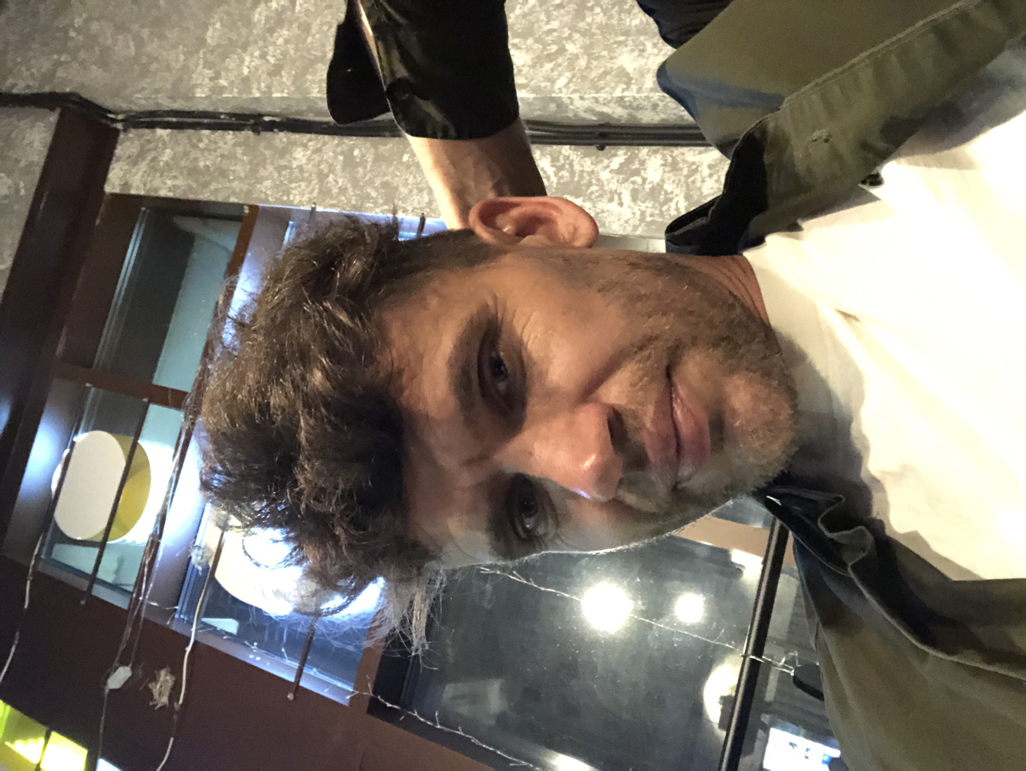 The image is a close-up selfie of a man with messy brown hair, a light beard, and a white t-shirt visible under a dark green jacket. He is looking directly at the camera with a neutral expression. His right arm is visible behind him, resting on his head, and he is wearing a black long-sleeved shirt or jacket. The background appears to be an indoor setting with a window, exposed wires, and textured walls. The lighting suggests it might be evening or a dimly lit interior. The man's clothing and the indoor setting do not provide specific clues to pinpoint the exact location within Saint Petersburg, Russia.