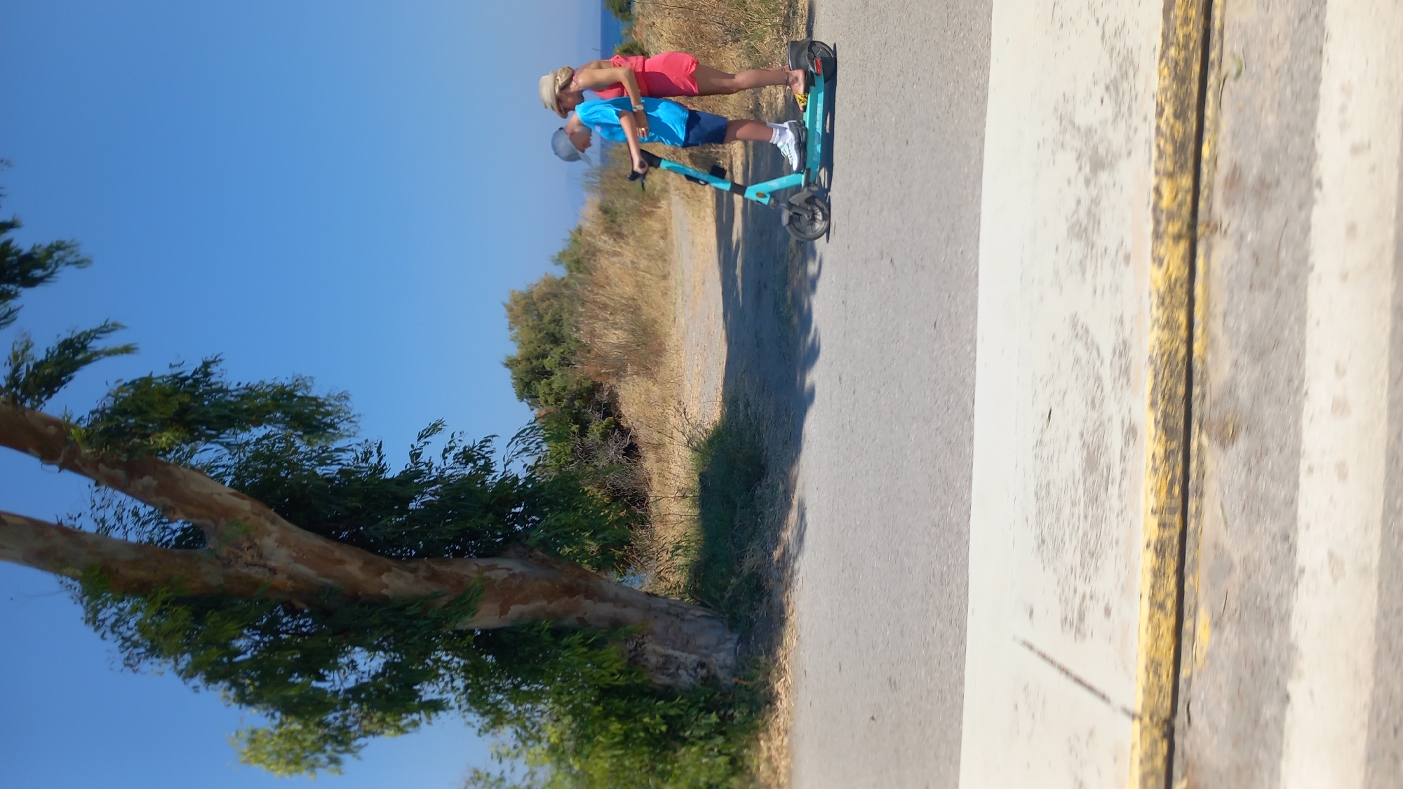 A boy and an adult woman are riding on a teal electric scooter on a paved path. The boy is in front, steering the scooter, and the woman is behind him, with her hands on his waist. Both are looking forward. The boy is wearing a blue t-shirt, dark shorts, a grey baseball cap, and white sneakers. The woman is wearing a bright pink dress and a straw hat. They are on a paved path that appears to be in a park or coastal area, with dry grass and bushes on either side. A large tree with a light-colored trunk and green leaves is to the left of the path, and the blue sky is visible above. The sun is shining brightly, casting shadows. In the foreground, there is a section of the paved path with yellow and white markings. The background shows more dry vegetation and a glimpse of the sea and sky in the distance.