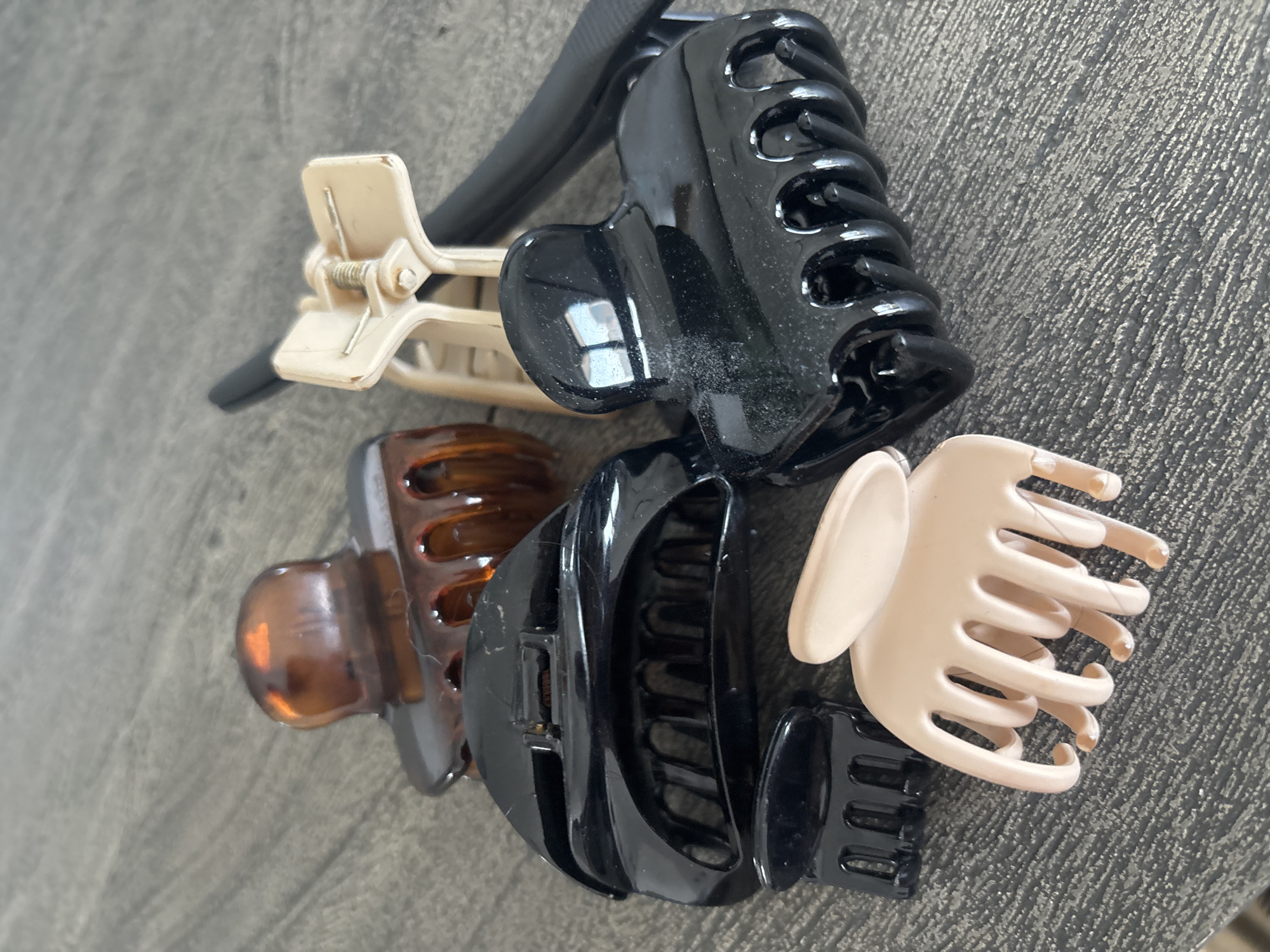 A collection of six plastic hair clips are arranged on a textured grey surface. The clips vary in color and size. One clip is a translucent amber, two are opaque black, one is a pale beige, and another is a slightly darker beige. A black hair accessory with a ridged texture is also partially visible. The image does not provide any information about the location in Dubai, United Arab Emirates.