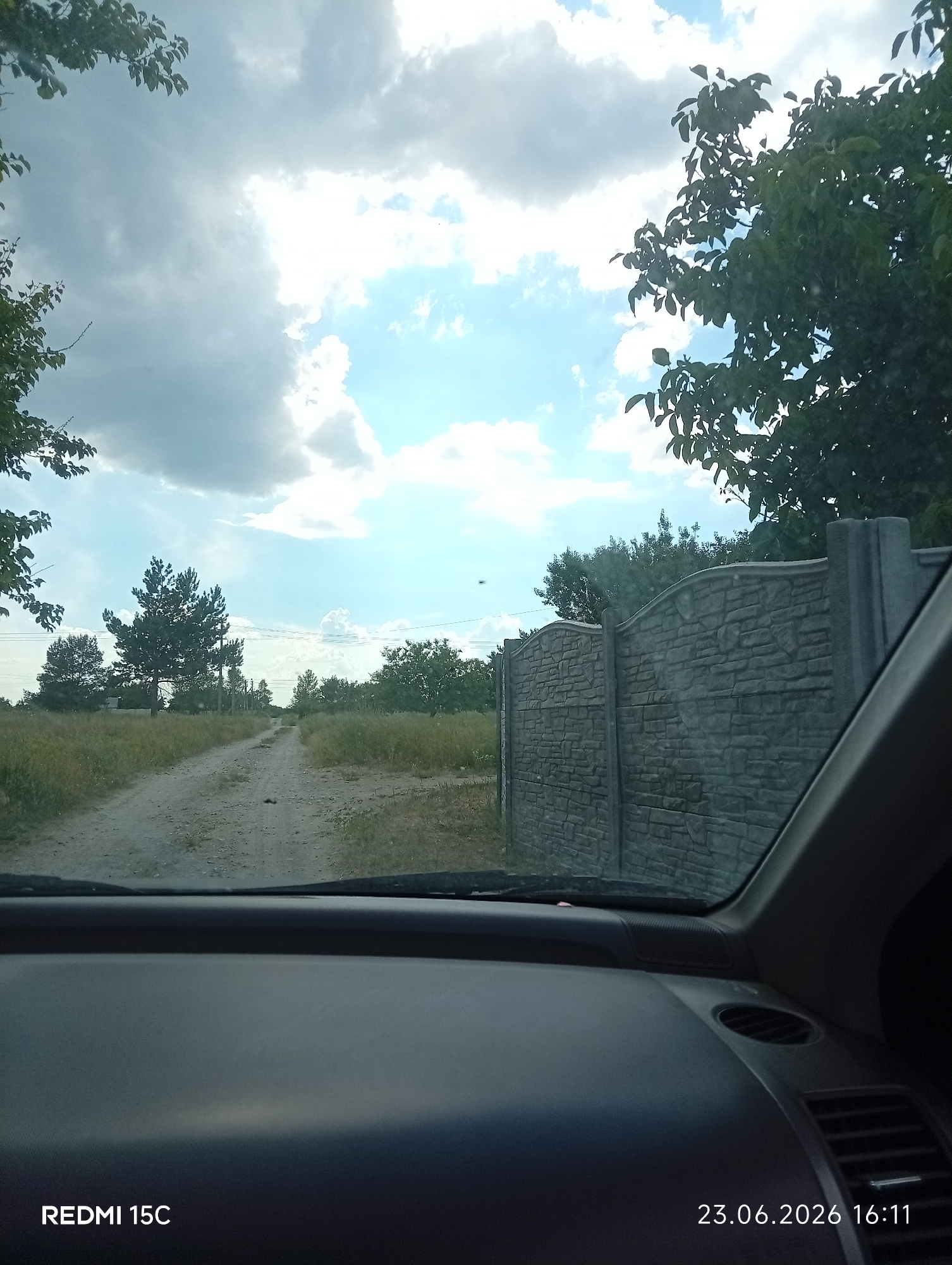 This is a first-person perspective photo taken from inside a vehicle, looking out at a rural scene. The foreground is dominated by the dark, textured dashboard of the car. Beyond the windshield, a dirt road stretches into the distance, lined with dry grass and scattered trees. The sky is partly cloudy with patches of blue visible, suggesting a pleasant, possibly overcast day. To the right, a stone-patterned fence runs along the edge of the property. A few trees with green foliage are visible in the middle and background, adding to the natural setting. The overall impression is one of a journey through a rural area.
Visible text includes "REDMI 15C" in the bottom left corner and a date and time stamp "23.06.2026 16:11" in the bottom right corner. The location is Mykolaivka, Ukraine.