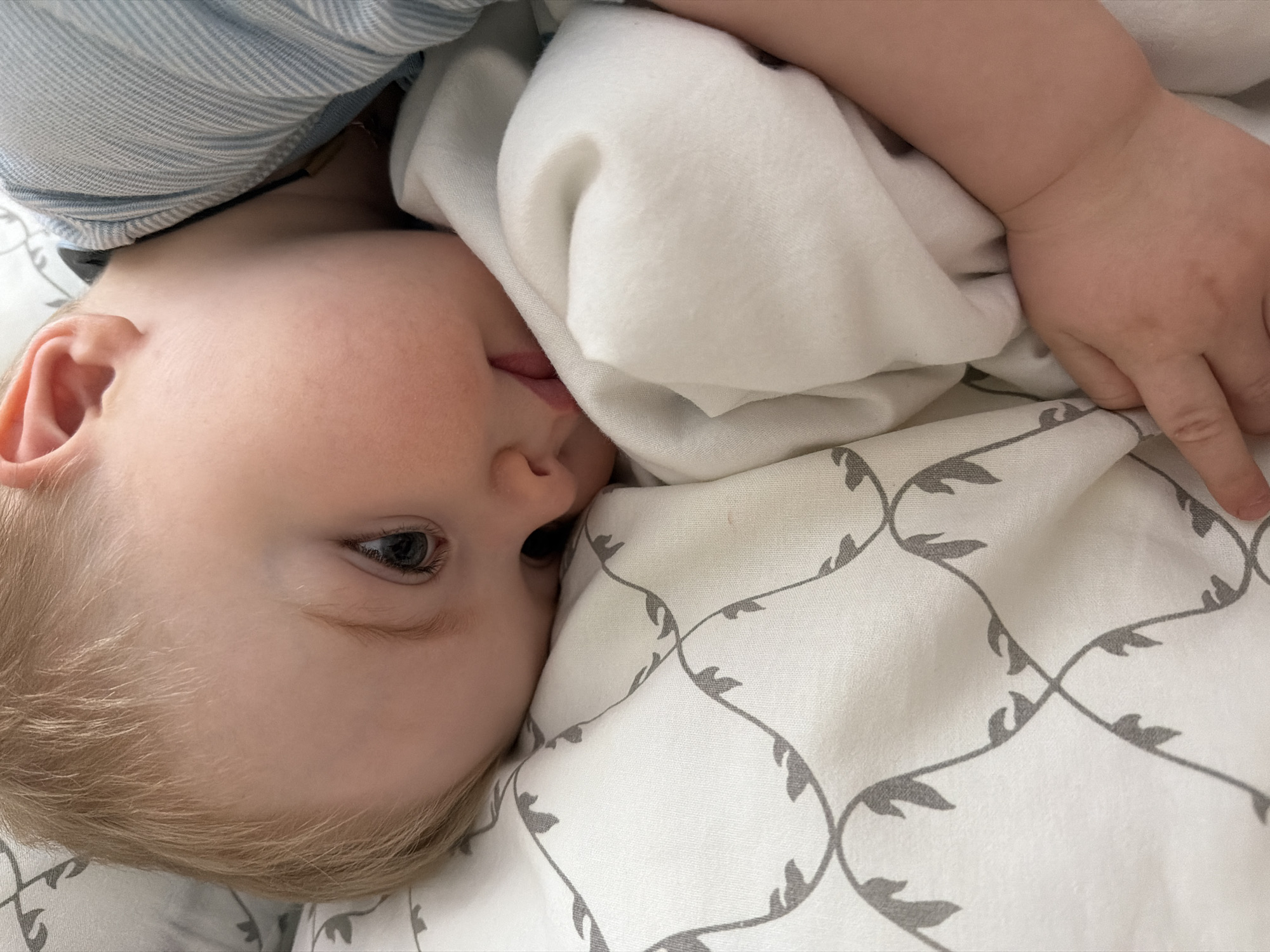 A baby with blond hair lies on a patterned white blanket, partially covered by a plain white blanket, with a light blue striped garment visible near the baby's head. The location is not determinable from the image.