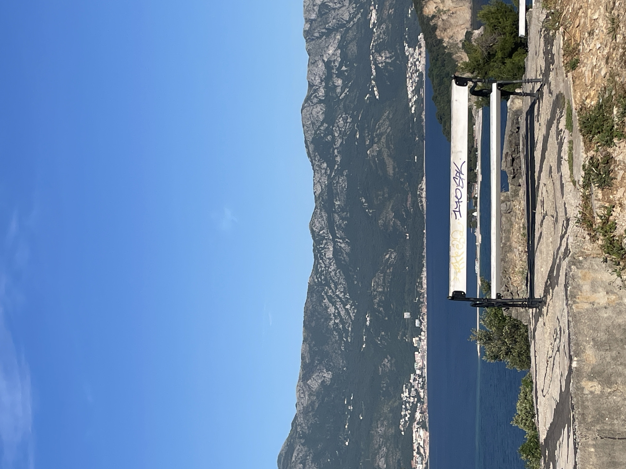 The image depicts a view overlooking the sea in Budva, Montenegro. A metal bench with graffiti is situated on a paved walkway along the edge of a cliff. In the background, a mountain range stretches along the coast, and the sea extends to the horizon under a clear blue sky. The buildings of Budva are visible nestled along the coastline at the foot of the mountains. The foreground shows weathered stone along the walkway and sparse vegetation growing along the cliff's edge.