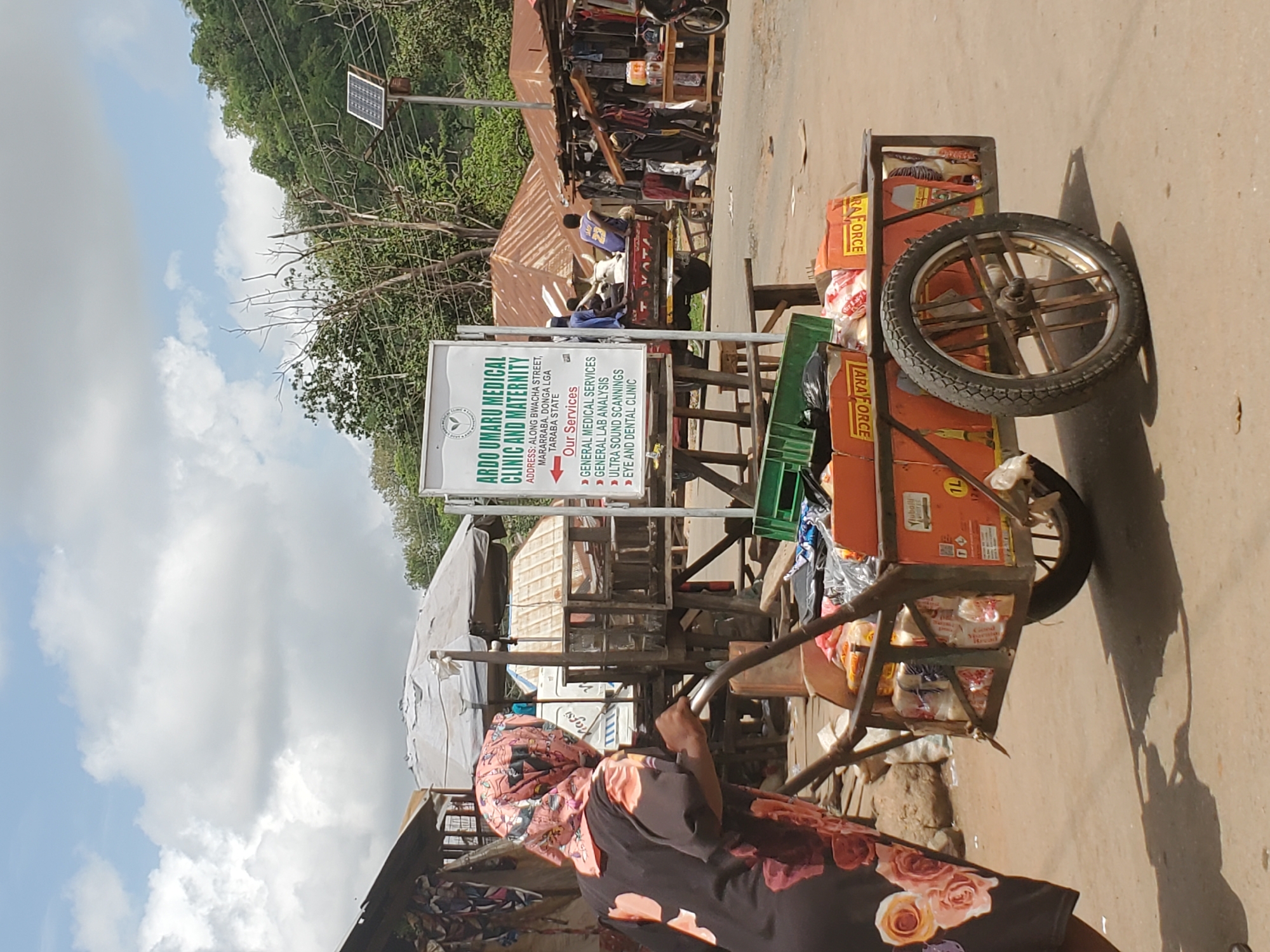 A woman wearing a floral print dress and head covering is pushing a metal cart loaded with goods down a paved street in Mararaba, Nigeria. The cart is stocked with boxes labeled "LARA FORCE," possibly laundry detergent, and other items. A sign for the "ARDO UMARU MEDICAL CLINIC AND MATERNITY" is visible in the background, indicating its location on Bwacha Street. Other small structures and people are present along the street, suggestive of a marketplace or commercial area. A small solar panel is mounted on a pole. The sky is partly cloudy.