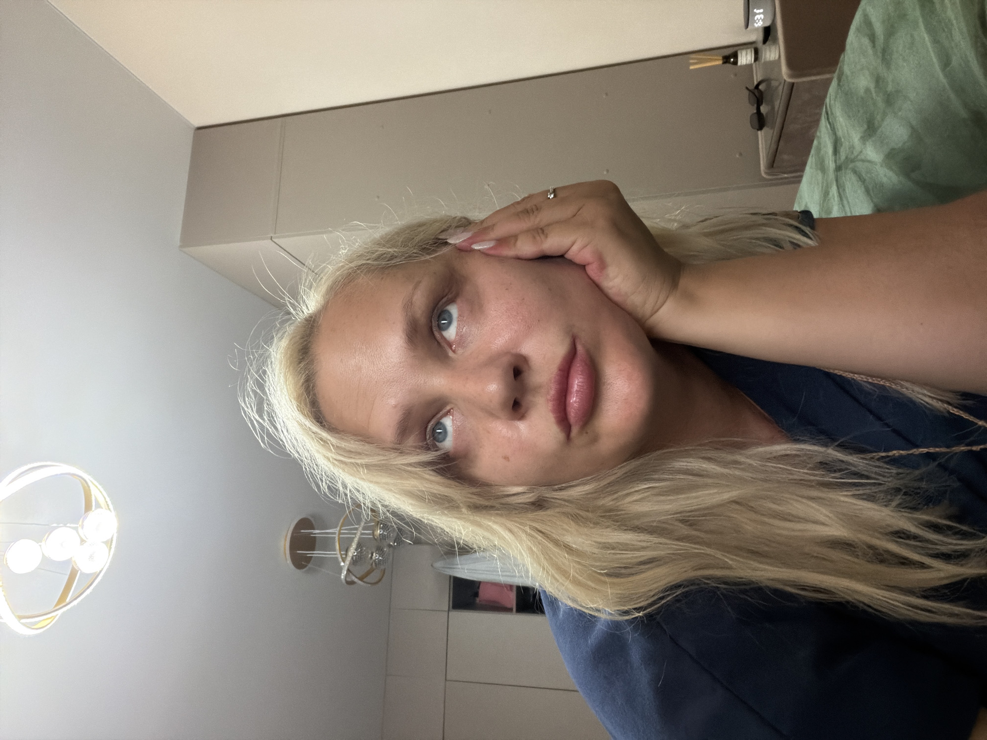 A blonde woman is lying down, resting her head on her hand, and looking upwards. She is wearing a dark blue top. The background includes a ceiling with two modern light fixtures, a wardrobe or closet, and a dresser with a small bottle of diffuser oil and sunglasses. The overall environment appears to be an indoor residential setting. There is no information available to determine the location in the city of Kazan, Russia.