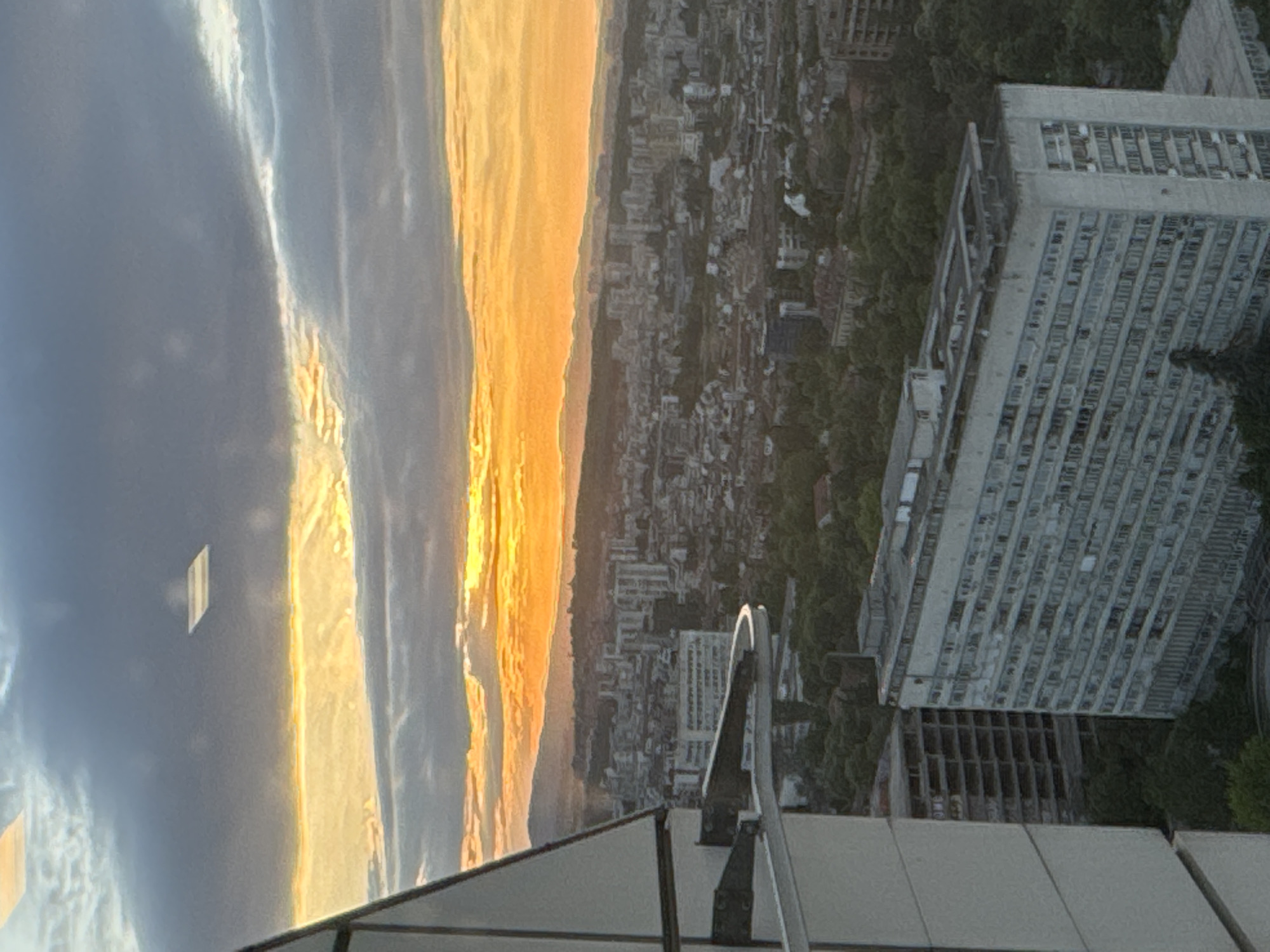 The image shows a cityscape at sunset. The sky is divided with a blue, cloud-filled side on the left and a bright golden-orange side on the right, suggesting a sunset. Below the sky is a dense urban area with various buildings and greenery, likely a city. A large, multi-story white building is visible prominently in the mid-right of the image. A balcony or rooftop structure is visible in the lower part of the image, suggesting the photo was taken from an elevated position within a building.
Given the location is Sofia, Bulgaria, the large, multi-story white building in the mid-right of the image is likely a building located in Sofia.