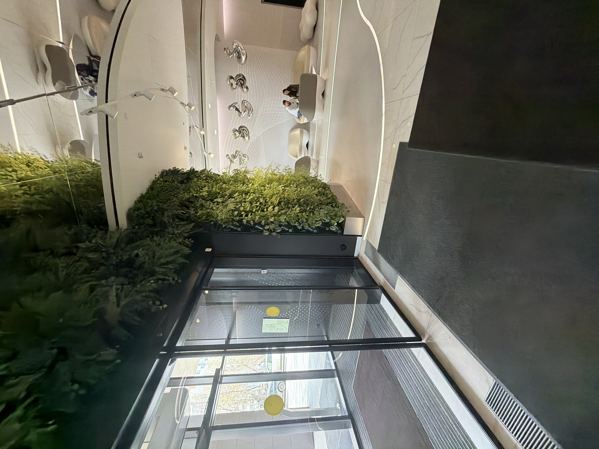 The image depicts an interior view of a building in Moscow, Russia. A dense, lush green plant arrangement is positioned near a floor-to-ceiling glass entrance, reflecting a street scene. The building's interior has a modern design, with a curved wall, decorative metal sculptures, and what appears to be a waiting area with people seated on stylized furniture. The architectural style is contemporary and polished, and the color palette is predominantly neutral, with white, gray, and black elements complemented by the natural greenery. The scene suggests a high-end commercial or residential space, well-maintained and thoughtfully designed.
